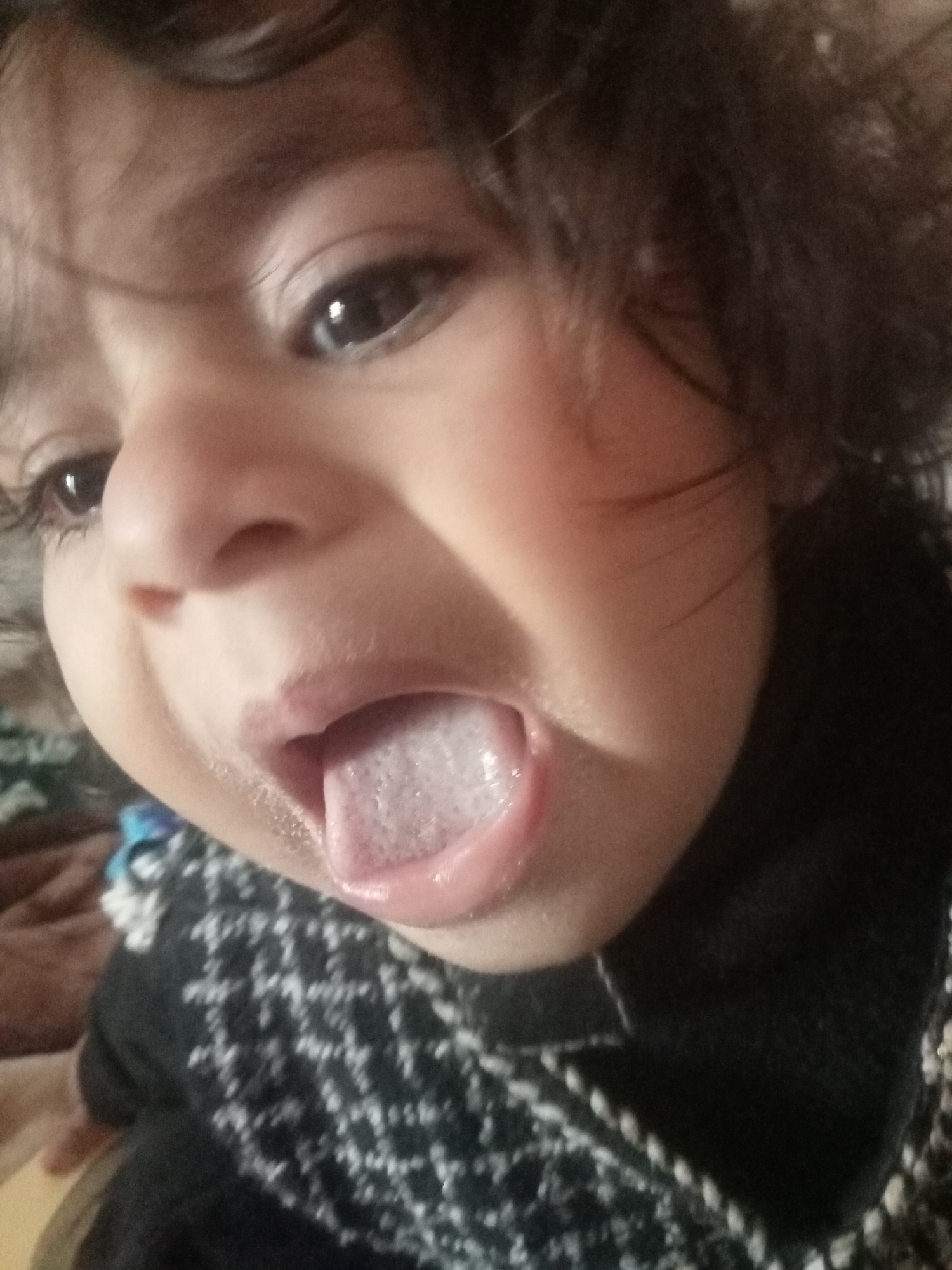 This is a close-up, slightly high-angle portrait of an infant or young child, located in Mansehra, Pakistan. The subject has dark, wide-open eyes looking upward and to the left, and dark, curly hair. The child's mouth is open, with the tongue extended and exhibiting a distinct whitish coating. The skin tone is light brown. The subject is wearing a dark-colored garment with a patterned knit design in white or a light contrasting color. The background is out of focus, showing blurred textures and colors consistent with an indoor environment. No other individuals or specific objects are clearly discernible in the scene.