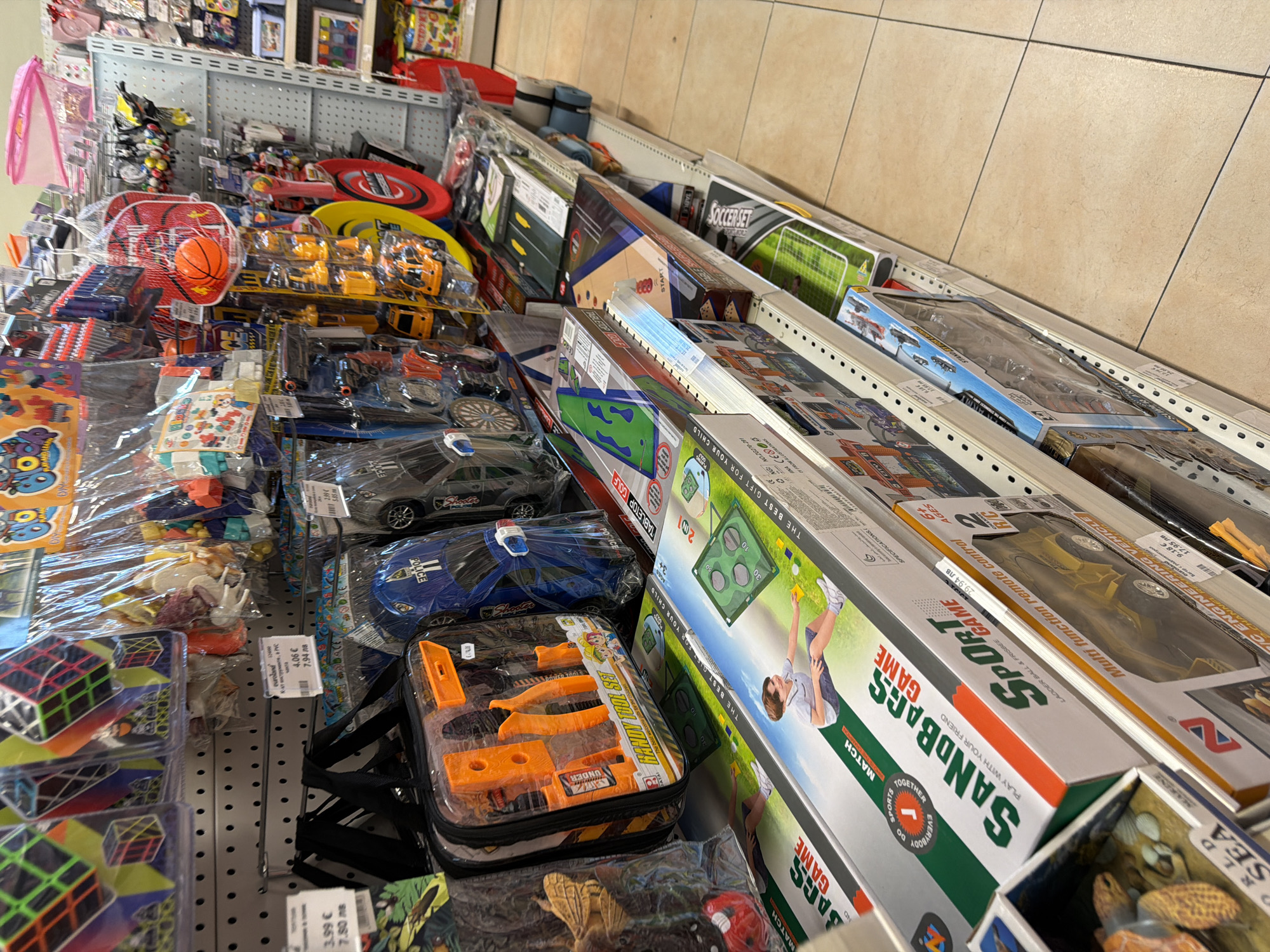 The image is a medium-angle, full-shot photograph of a retail store aisle, likely a toy store, filled with various toys displayed on shelves. The shelves are made of white perforated metal, and the flooring consists of beige tiles. The overall lighting is bright and even.
In the foreground, there are several toy boxes stacked on shelves. The most prominent box, positioned slightly to the left of center, is labeled "SaNDBaGS SPORt GAME". It depicts a young boy playing a game on a green field with a yellow ball and a target. To the right of this box, another toy box is visible, featuring a yellow remote-controlled vehicle labeled "Multi function remote control". Below these, a toy with a turtle-like design and the text "WORLD SEA" is partially visible.
Further up the shelves, a clear plastic bag contains an orange toy tool set, including pliers and other tools, with the label "HANDY TOOL SET". Adjacent to this, a blue toy car with "POLICE" written on it is displayed.
In the upper portion of the image, more toys are visible, including colorful Rubik's cubes in plastic packaging, bags of small figures, and a red basketball hoop with an orange basketball. Other merchandise, such as plastic baskets and packaged games, are also present on the shelves.
The image does not contain any identifiable individuals. The scene is a static retail display of merchandise.
Based on the provided price tags, one of which is in Bulgarian (e.g., "4,06 лв 7,94 лв"), this retail store is likely located in Pazardzhik, Bulgaria. The price format and the Bulgarian currency abbreviation "лв" (leva) strongly suggest this location.
