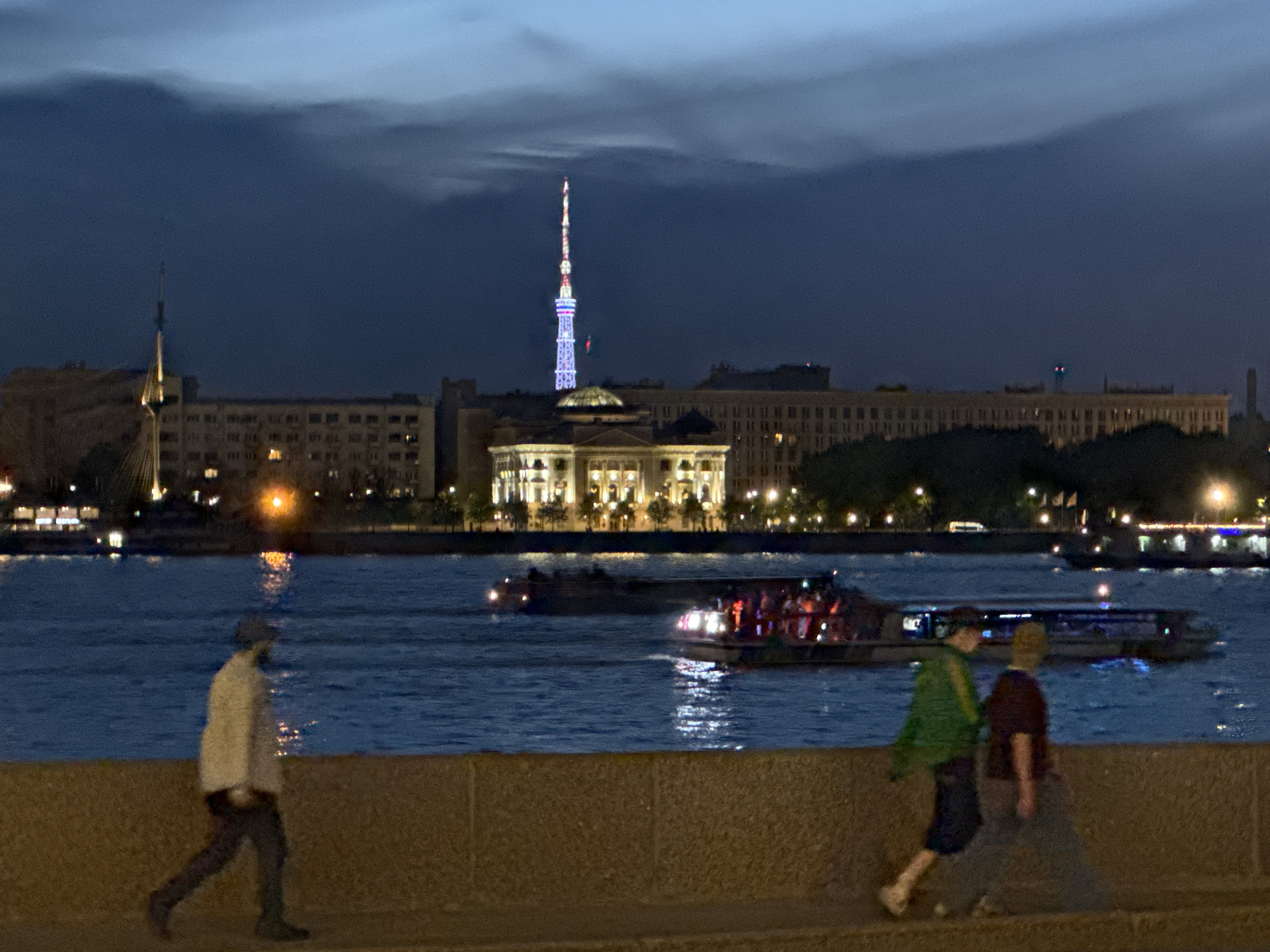 This nighttime image captures a panoramic view of the Neva River embankment in Saint Petersburg, Russia, featuring the illuminated Saint Petersburg TV Tower and surrounding city architecture under a dark, cloudy sky.
In the foreground, a rough-textured stone embankment or wall spans the width of the frame. Three indistinct individuals walk from left to right on the pedestrian path alongside the wall. The leftmost person wears a light-colored jacket, dark pants, and a dark cap. To their right, two individuals walk in close proximity; one wears a green jacket and dark lower garments, while the other wears a dark reddish-brown top and lighter, possibly grey, lower garments. Their forms are blurred due to motion or low light conditions.
The midground is dominated by the dark blue waters of the Neva River, reflecting numerous bright white and yellow lights from the city and boats. Several vessels traverse the river. A prominent passenger boat, illuminated with white and reddish lights and showing multiple indistinct figures on deck, moves from left to right in the center-right. Another boat is partially visible behind it. Further to the left, another darker vessel is present.
The background showcases the cityscape of Saint Petersburg. The most prominent feature is the brightly lit Saint Petersburg TV Tower, positioned centrally, displaying alternating red, white, and blue sections. Below the tower, a large, classically styled building with a central dome and numerous illuminated windows stands out. To the left and right of this central structure, long multi-story residential or commercial buildings with many lit windows line the riverbank. On the far left, a tall, slender mast-like structure with a yellow glow is visible. Scattered trees and foliage are present along the river's edge in front of the buildings. The overall scene is lit by artificial city lights against a twilight or nighttime sky.