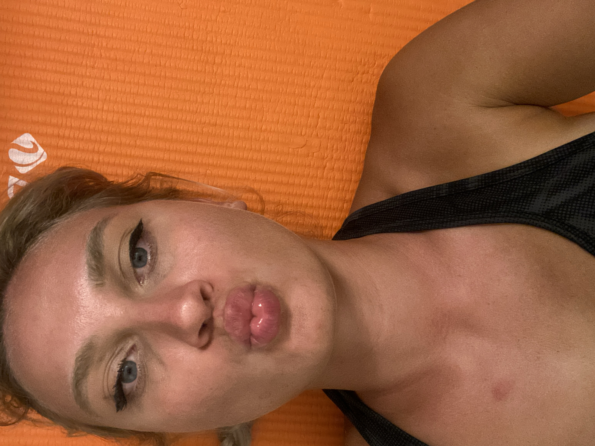A young woman with blonde hair, blue eyes, and visible makeup is captured in a close-up, selfie-style photograph. She is making a kissing face, with her lips puckered. She is wearing a dark-colored top with a halter neckline. The background is an orange, textured surface, possibly a yoga mat or similar item. A small, reddish mark is visible on her upper chest. The lighting suggests an indoor setting. The image does not provide any specific information or context that would allow for its location to be determined within the city of Aksu, Turkey.
