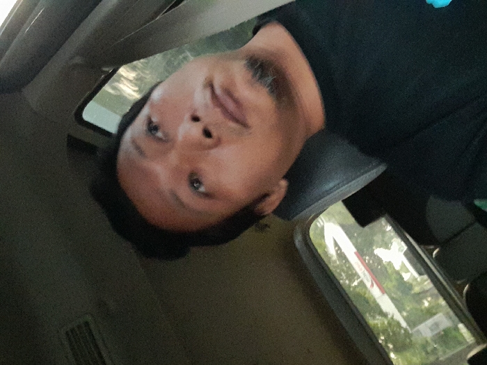 A single East/Southeast Asian male is depicted seated in the interior of a vehicle within the city of Temuwulan, Indonesia. The individual has short dark hair and a goatee, and is wearing a dark t-shirt. His head is tilted slightly upwards and to the right, with his gaze directed off-frame towards the upper right. The subject's head rests against a dark upholstered headrest. The vehicle's dark interior ceiling, an overhead air vent, and window frames are visible. Through the vehicle's rear side window, an exterior daytime environment is observed, featuring green foliage and a partially obscured white and red horizontal structure, consistent with a barrier or sign. No explicit actions or interactions are occurring within the frame.