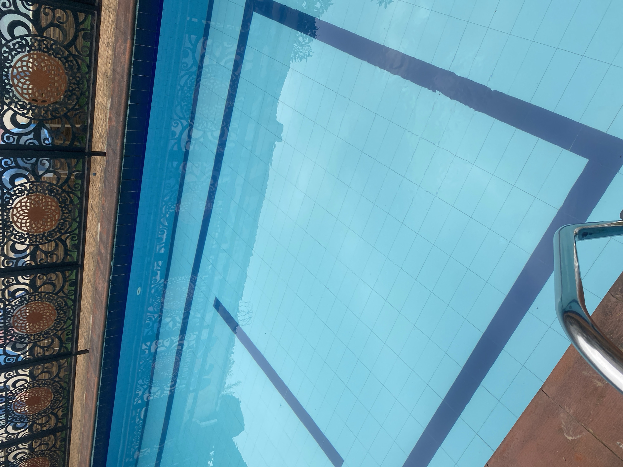 This image shows a swimming pool, likely located in Ikorodu, Nigeria. The pool is lined with light blue tiles and features dark blue lane markers. The water surface reflects the sky and possibly surrounding structures. A metal railing is visible at the pool's edge, suggesting an entry or exit point. A decorative metal fence is present along one side of the pool, next to what appears to be a tiled or brick surface. No people are visible. The environment suggests an outdoor recreational space.
