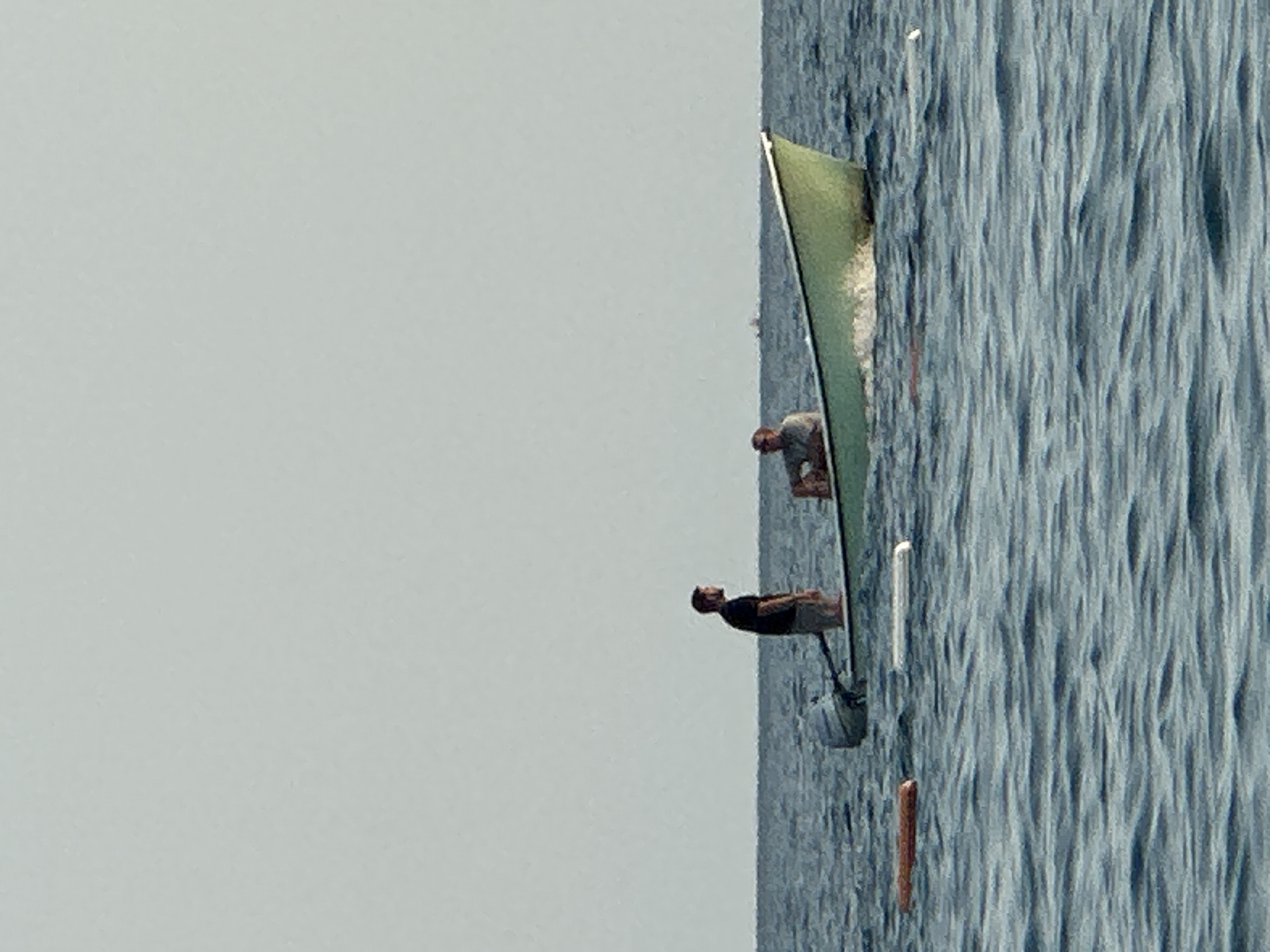 In the city of Boreti, Montenegro, two individuals are in a small, light green boat navigating the calm, blue water of the sea. The boat is equipped with a small outboard motor. One person, wearing a dark t-shirt and shorts, stands at the rear of the boat, facing away from the viewer. The second person, wearing a light-colored shirt, is seated towards the front of the boat, also facing away from the viewer. The sky is a uniform, pale grey, suggesting an overcast day or early morning/late evening light. Several buoys are visible in the water: one reddish-brown buoy is to the left of the boat, and a white buoy is located to the right. The water exhibits gentle ripples. In the distance, a very small, dark silhouette of what appears to be another boat or structure is faintly visible on the horizon.