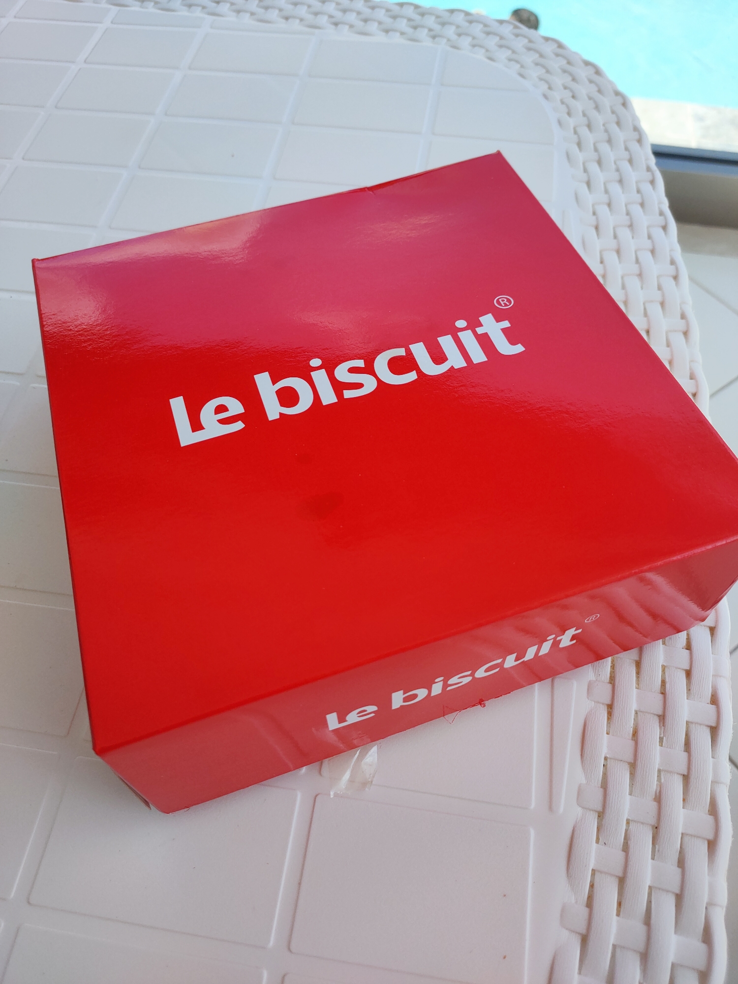 A red box with "Le biscuit" printed in white text rests on a white, textured surface. The box is positioned diagonally, with the top-left corner closer to the viewer. Part of a white, woven surface, possibly a chair or table, is visible to the right of the box. In the background, a blurry expanse of blue water and a railing suggest an outdoor setting, possibly a swimming pool area. The location is identified as Mrezga, Tunisia.