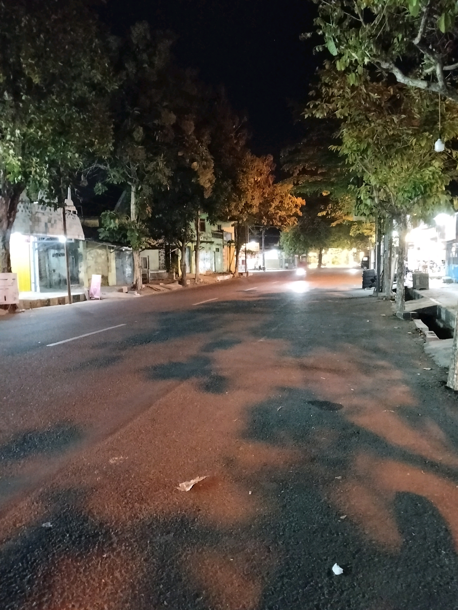 A wide asphalt road is depicted at night in Purworejo, Indonesia, with streetlights casting a warm glow on the wet pavement. Trees line the sides of the road, their leaves illuminated by the artificial light. Buildings, appearing to be shops or residences, are visible in the background, some with lights on. A motorcycle is seen in the distance, its headlights creating a bright streak on the road. There are no people clearly visible in the scene, though shadows and the presence of the motorcycle suggest activity. The overall atmosphere is calm and quiet, characteristic of a residential or commercial street at night.
