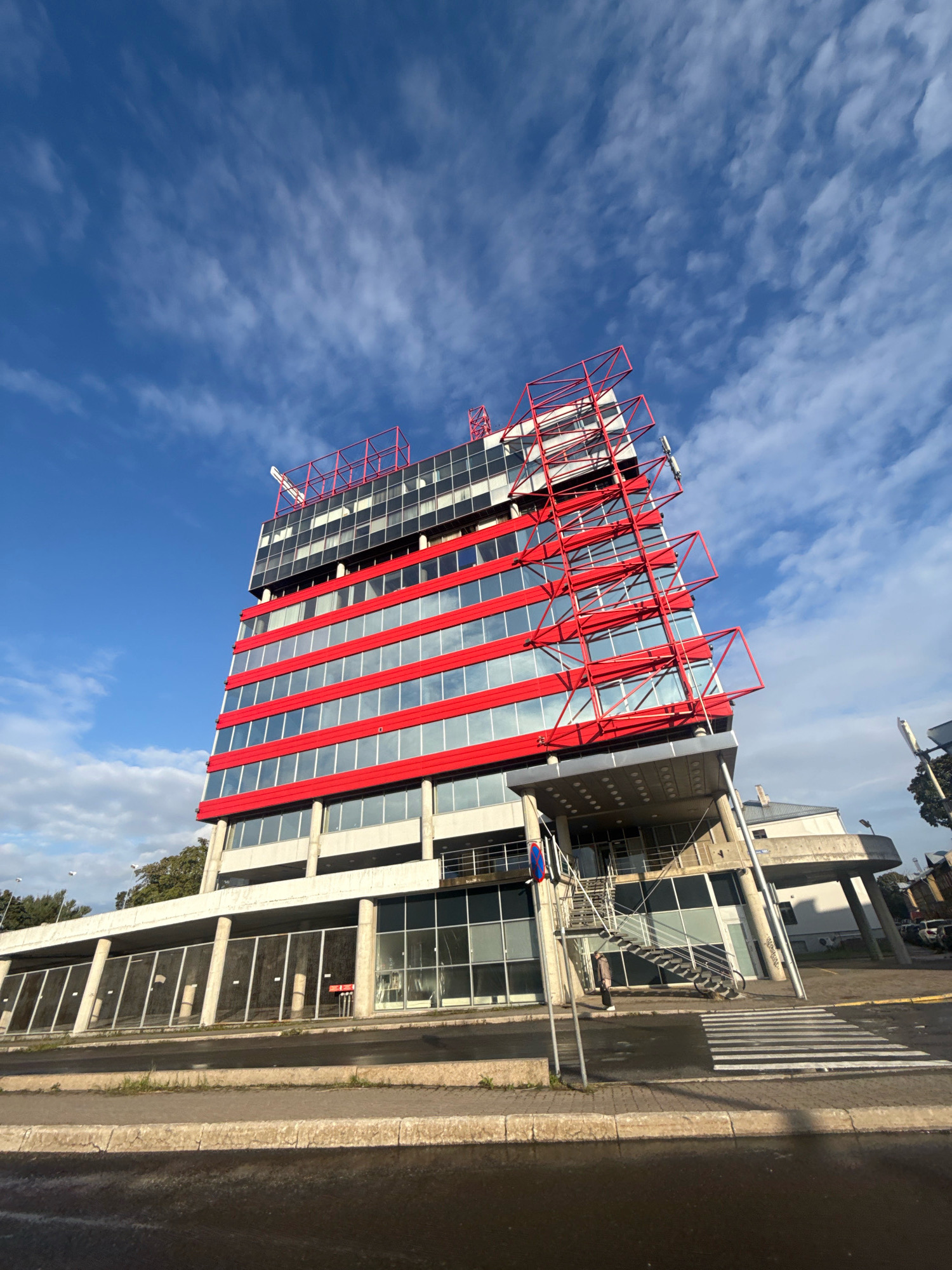A low-angle shot captures a modern, multi-story building with a striking red and glass facade against a bright blue sky with scattered white clouds. The building features horizontal red bands interspersed with rows of reflective glass windows. A prominent red metal framework extends upwards from the top of the building, resembling an abstract sculpture or scaffolding.
In the foreground, a wet street and sidewalk are visible, with a pedestrian crossing marked by white stripes. The base of the building has large windows and pillars. A set of metal stairs leads up to an elevated entrance on the right side of the building, where a person in dark clothing is descending. The overall impression is one of contemporary architecture situated in an urban environment.
The building appears to be located in Tallinn, Estonia, likely in an area with modern commercial or office spaces. The architecture suggests a relatively new development. The wetness of the street indicates recent rainfall.