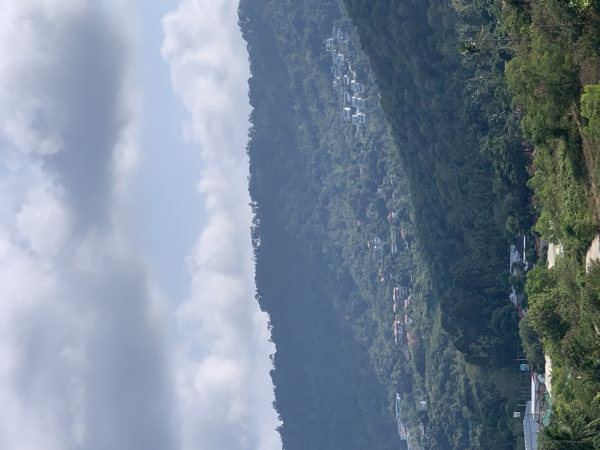 The image depicts a mountainous landscape in the บ้านบางรักษ์ area of Thailand. The mountain is densely vegetated with various trees and plant life. Several buildings are scattered along the mountainside, varying in size and appearance. The sky is visible on the left, filled with white and gray clouds. The picture is oriented in a portrait style, but turned on its side.