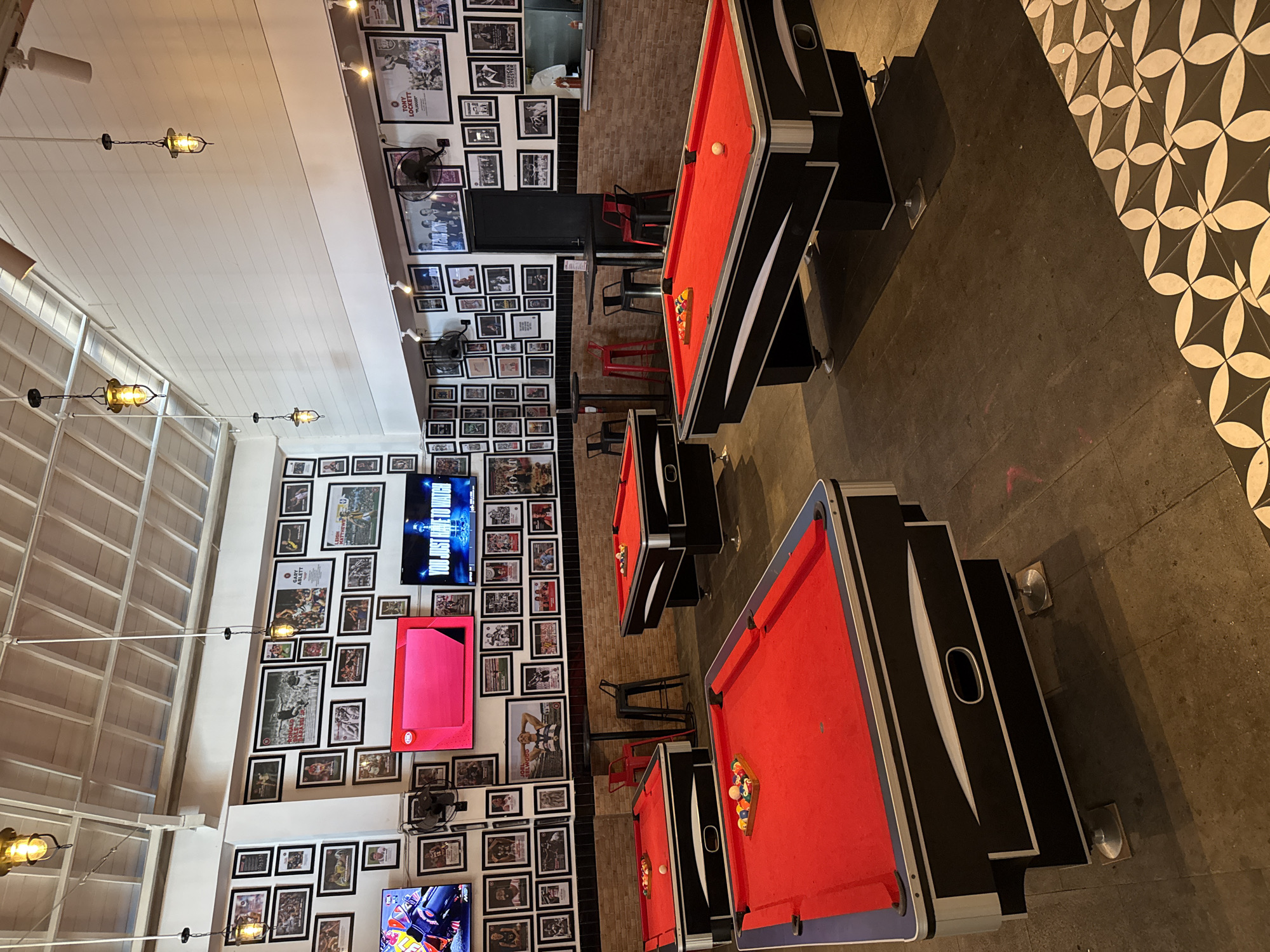 The image captures the interior of a dimly lit entertainment venue, likely a bar or billiards hall. The primary subjects are multiple pool tables, each featuring red felt playing surfaces and black frames with silver accents. Some tables have billiard balls racked or scattered on them.
The walls are densely covered from floor to ceiling with numerous framed pictures and memorabilia, primarily photographs, many appearing to be sports-related, in both black and white and color. Several flat-screen televisions are mounted on the walls, displaying various content; one screen clearly shows the phrase "YOU JUST HAVE TO WATCH." Small wall-mounted fans are also visible on the picture-covered walls.
The ceiling is white and paneled, with several industrial-style pendant lights hanging down, casting a warm glow. The floor is dark, appearing to be concrete in most areas, with a section on the right featuring decorative black and white geometric patterned tiles. In the background, there are high-top tables and red and black bar stools, alongside a section of exposed brick wall. No people are visible in the scene.
Visible text on the framed pictures includes "TONY LOCKETT", "GARY ABLETT", and "JOEL SELWOOD", suggesting a sports theme. The overall atmosphere suggests a casual place for playing pool, watching sports, and socializing, likely in the evening.