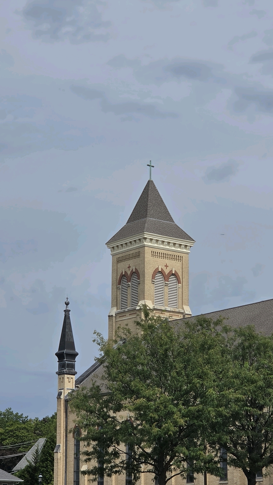 A medium shot shows the upper portions of a tan brick church and a smaller, darker steeple against a cloudy sky. The main church tower has a gray shingled roof topped with a green cross. Two arched windows with louvers are visible on the tower, each framed by red brick. A smaller, dark steeple with a finial is visible to the left of the main tower. Dense green trees obscure the lower part of the church building. The scene is set in East Dundee, United States of America.