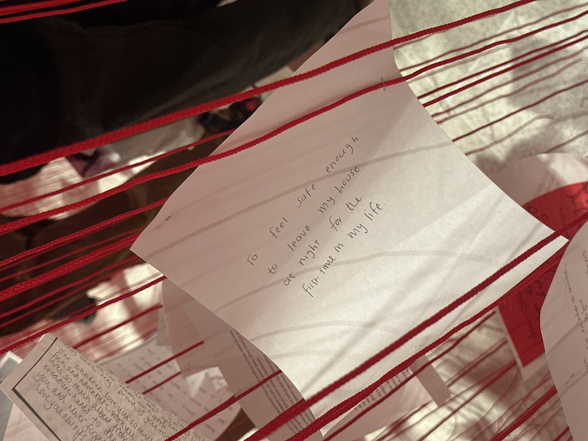 The image shows pieces of paper suspended by red strings. The papers contain handwritten text. One paper says "To feel safe enough to leave my house at night for the first time in my life". Another paper reads "Our children love you so much fun and adventure with you lives so much album from treasure hunts to you and never forget Love you always." The background is blurred, suggesting a shallow depth of field. The environment includes a bed or similar surface with striped linens.