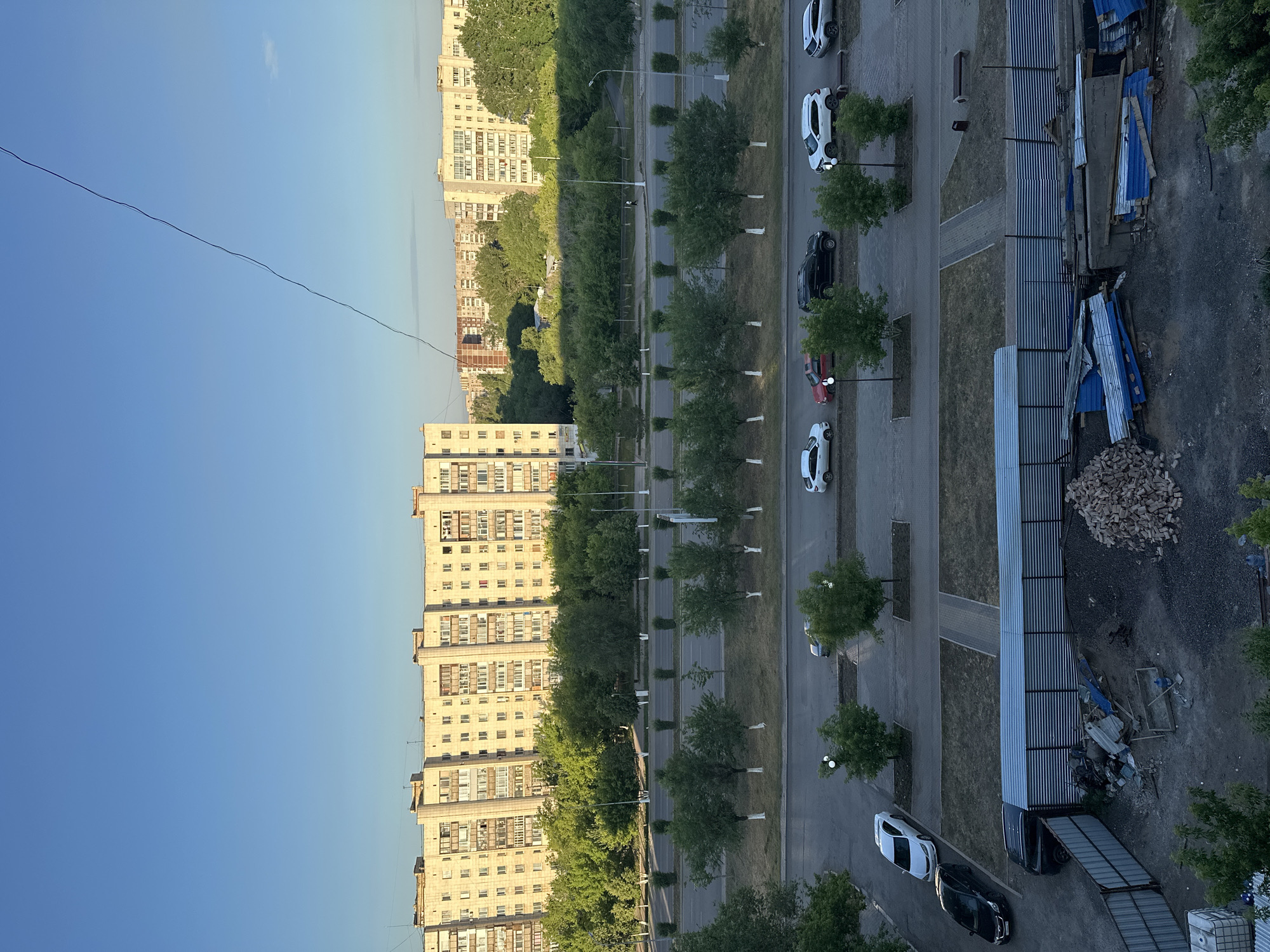 The image captures an urban landscape in Karaganda, Kazakhstan, under a clear blue sky. In the background, a long, multi-story apartment building with a beige facade stretches horizontally. In front of the building, a dense line of green trees creates a visual barrier. A road runs parallel to the trees, with several cars parked and driving on it. Further down, another apartment building is visible, also with a light-colored facade.
In the midground, a street is lined with trees, their trunks painted white at the base, and lampposts. Cars are seen parked on this street and on a sidewalk area.
The foreground shows a construction site with fencing, piles of bricks, rubble, and building materials, including corrugated metal sheets and wooden planks. Several cars are parked in this area as well, including a black SUV and a white sedan. The ground is a mixture of dirt, gravel, and scattered debris. The overall scene depicts a residential area undergoing some form of construction or renovation.