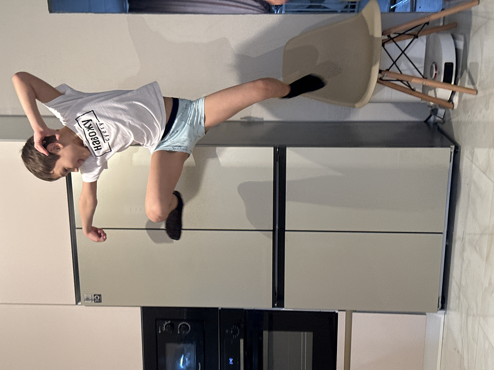 This image captures a dynamic scene featuring a young boy in an active pose within a modern kitchen setting in Moscow, Russia. The boy is depicted mid-air, horizontally suspended, suggesting he is jumping, falling, or performing a gymnastic move. He wears a white t-shirt and light blue shorts or boxers, with black socks on his feet.
His body is positioned with his head turned towards the left, his right arm bent with his hand near his head, and his left arm extended downwards. One leg is bent upwards, while the other is extended straight, with its foot seemingly near a beige chair. The chair, with its wooden legs and black metal frame, stands upright to the right of the boy. A white robot vacuum cleaner is partially visible on the floor near the chair's base.
The boy's white t-shirt has prominent black text. The main text is in Cyrillic: "навожу суету," which translates to "I'm stirring up a fuss" or "I'm making a commotion." Below this, smaller Japanese characters "すぅと" are also visible. His expression appears focused, possibly conveying effort or enjoyment.
The background reveals a contemporary kitchen environment. To the left are light-colored cabinets or a refrigerator panel, and to the right, a large, two-door refrigerator or integrated tall cabinets. Below these, black built-in ovens or microwaves are visible. The walls are light, and the overall lighting suggests it is daytime indoors.