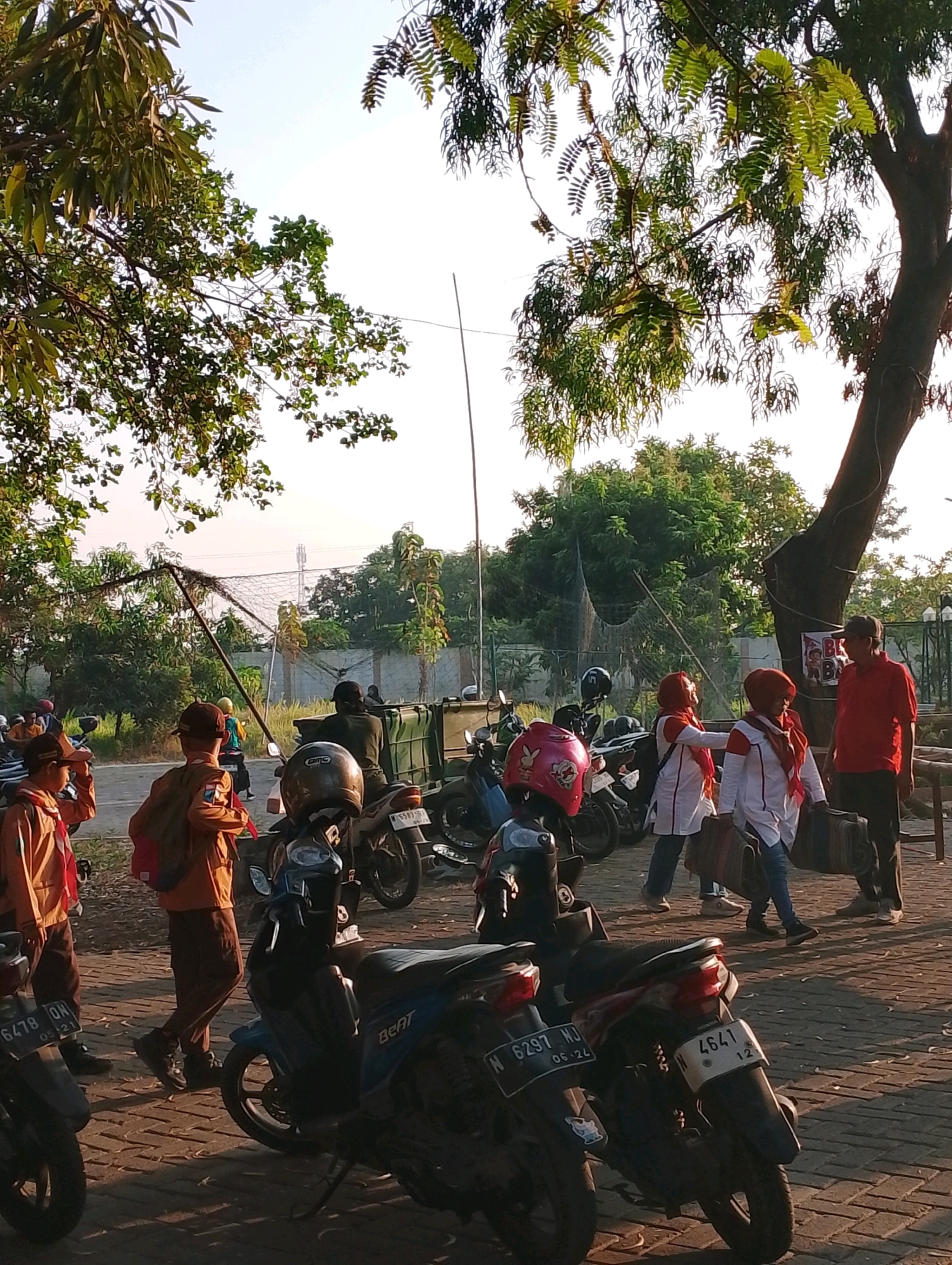 The image shows a group of people, some of whom appear to be students in uniforms, gathered around parked motorcycles. The scene appears to be outdoors, possibly a parking lot or a street in Probolinggo, Indonesia, given the presence of a license plate with the "N" prefix which is used for vehicles in East Java. Several students in brown and orange uniforms, some with backpacks, are visible in the foreground and middle ground. Some are looking towards the camera or interacting with each other. A man in a red shirt and a cap stands to the right, talking with two women wearing red headscarves and white tops. The environment includes trees with green leaves providing shade, a brick-paved ground, and a wire fence in the background. Several motorcycles are parked, some with helmets on them. The lighting suggests it is daytime, with the sun casting long shadows.