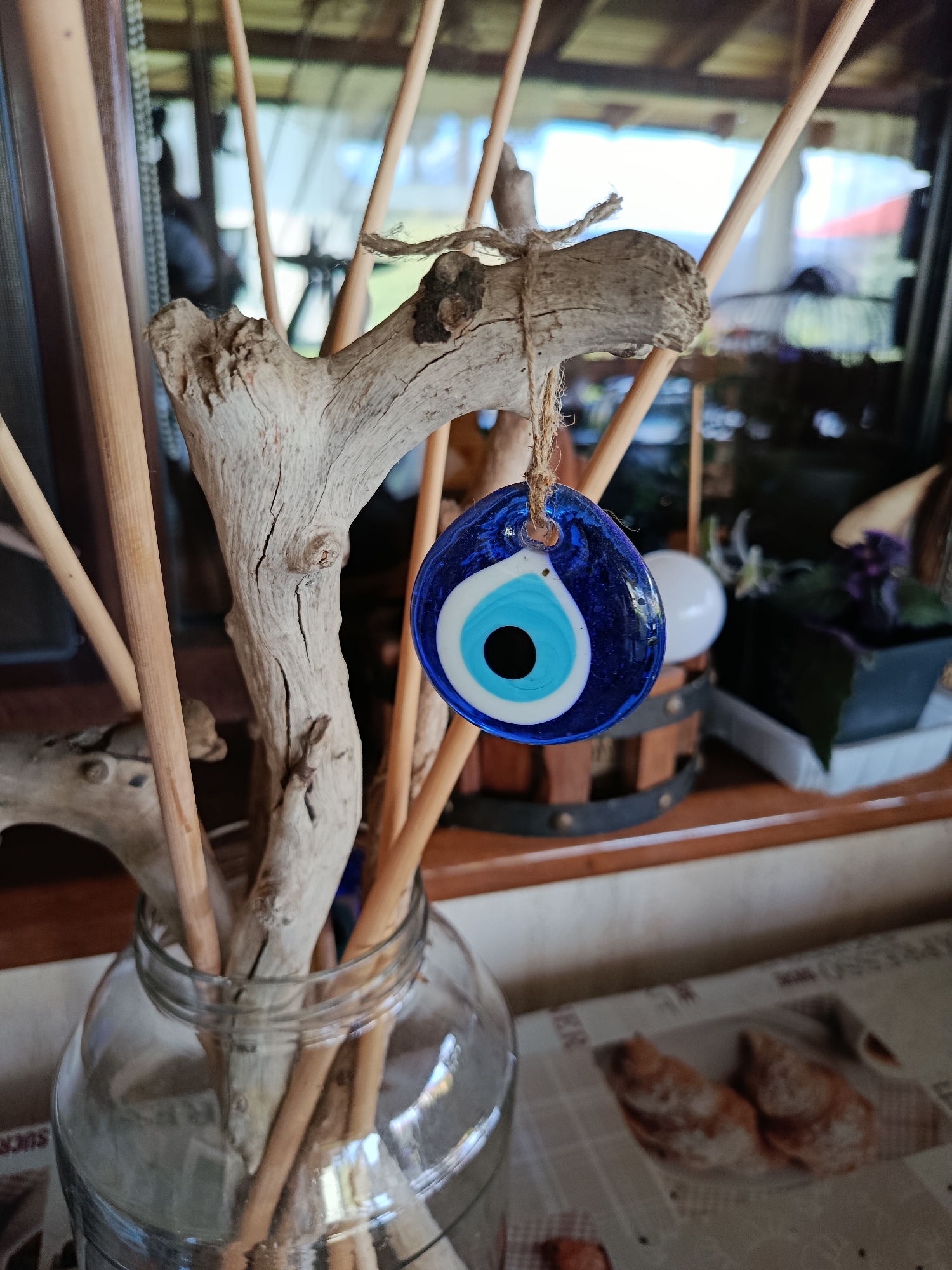 A close-up, eye-level, static shot captures a traditional blue and white evil eye amulet hanging from a piece of driftwood. The amulet, made of glass, features concentric circles of blue, white, and black, resembling an eye. The driftwood, weathered and grey, is adorned with a rough, bark-like texture and has several cracks. It is suspended by a thin, jute string. The driftwood itself is part of a larger arrangement of similar light-colored wooden sticks, some thin and smooth, others thicker and rougher, all emerging from a clear glass jar. The jar is filled with water, and the sticks are submerged in it, creating a natural, organic display.
In the background, a blurred scene suggests an indoor setting. A window or glass pane reflects an outdoor environment, hinting at greenery and possibly buildings. The overall ambiance is rustic and decorative, with elements of nature incorporated into the decor. The surface beneath the jar and driftwood appears to be a table, with a newspaper or printed material featuring images of bread visible in the lower right portion of the frame. The colors are muted, dominated by the natural tones of wood, glass, and the blues and whites of the amulet. There are no people or animals visible in the image. The scene is static and appears to be a still life arrangement.
The specific location in the city of Misaylovo, Russia, cannot be determined from the visual information provided in the image. The image depicts a decorative arrangement commonly found in various settings, but offers no geographical markers specific to Misaylovo or any other location.
