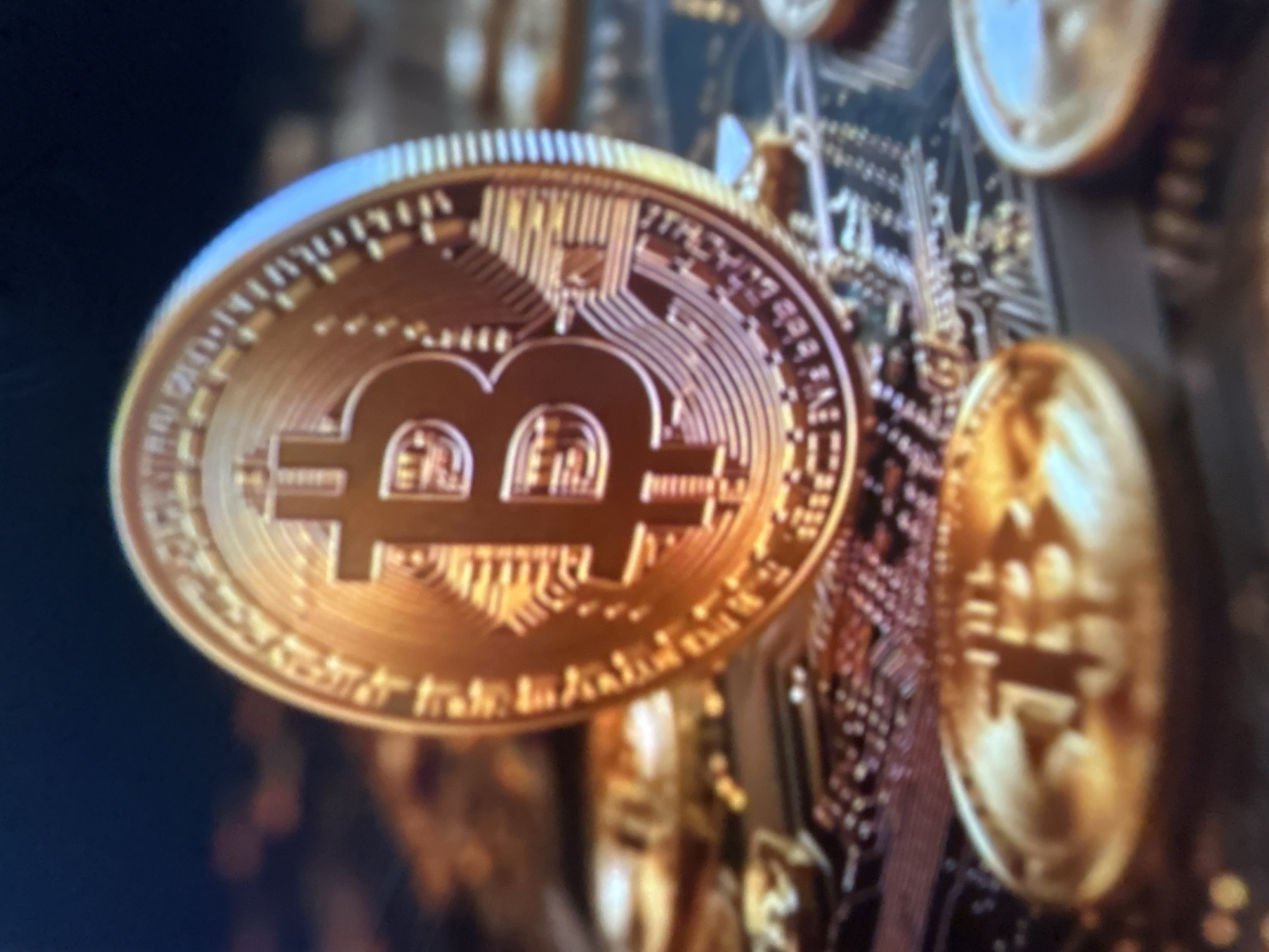 This media file depicts a close-up, slightly blurred shot of a gold-colored Bitcoin coin prominently featured in the foreground. The coin has the distinctive Bitcoin symbol "₿" embossed on it, along with intricate patterns and what appears to be text encircling its edge. Behind the primary coin, other similar coins and a blurry background suggestive of electronic circuitry or a digital landscape are visible. The lighting is warm and golden, highlighting the metallic sheen of the coins. There is no discernible activity or event taking place; the image appears to be a still representation focused on the object itself. The setting is abstract and digital, with no specific location cues or temporal indicators like time of day or weather. No emotions are conveyed, as the subject is inanimate. Any text on the coins is too blurred to be legible. The location context provided, Gelendzhik, Russia, is not visually represented in the image.