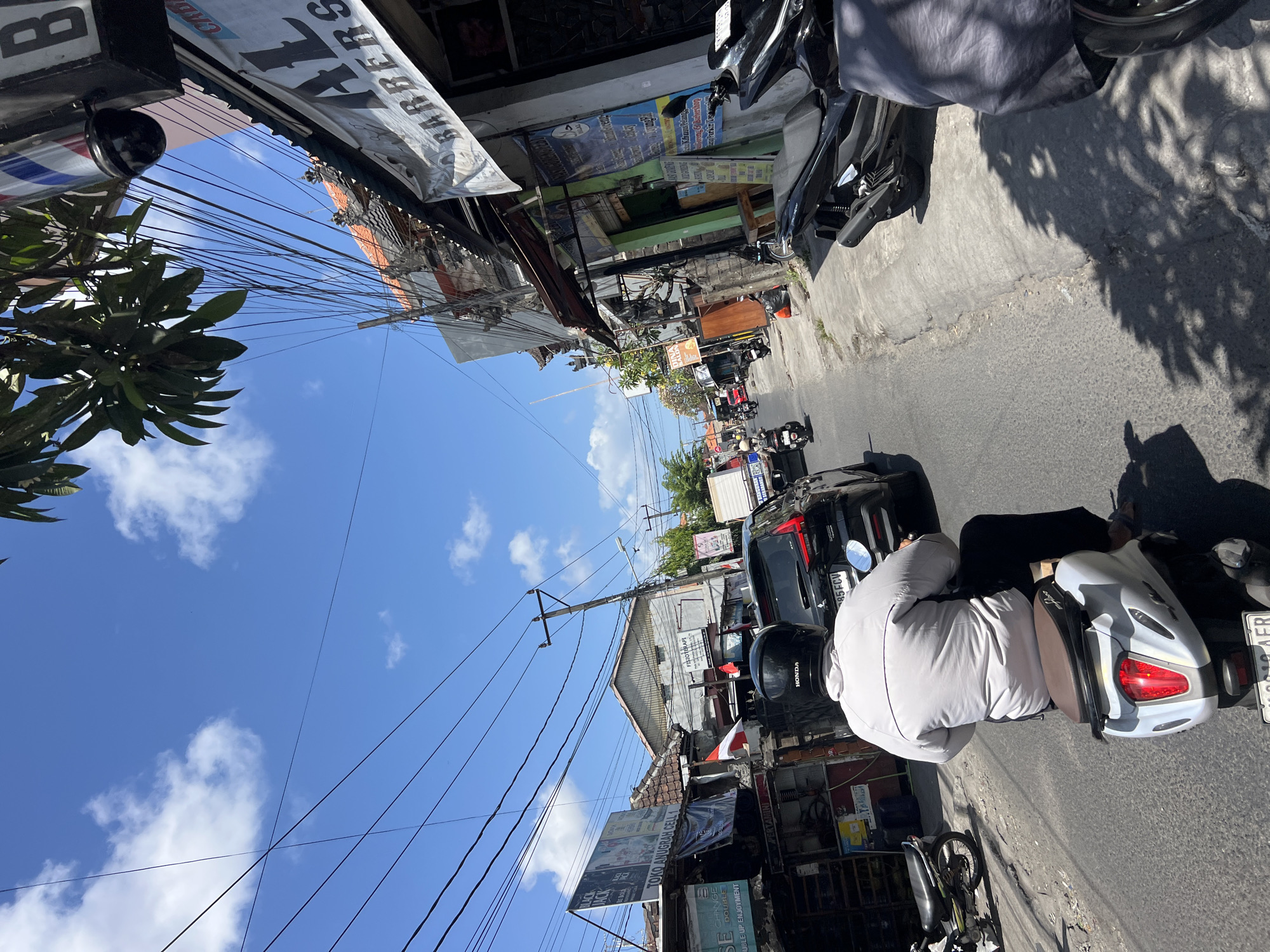 This is a street-level, eye-level, medium shot photograph taken outdoors in Denpasar, Indonesia, on a clear, sunny day with scattered white clouds. The image depicts a busy street scene with traffic, shops, and overhead power lines.
In the foreground, a person is riding a white scooter, viewed from behind. The rider is wearing a light-colored jacket and a black helmet. The scooter has a red taillight and a license plate that is partially visible.
To the right of the scooter, a black scooter is parked, partially covered by a grey fabric. Further down the street, other motorcycles and a black car are visible, indicating ongoing traffic.
On the right side of the street, a row of shops lines the road. One shop has a prominent sign that reads "AL BARBERS" with a barber pole above it. Other shop signs, some with Indonesian text, are also visible.
On the left side of the street, more shops and buildings are present, with some displaying advertisements and flags. A billboard with "TOKO ANUGRAH CELL 1" is visible.
Overhead, a dense network of electrical and telecommunication wires crisscrosses the sky, a common sight in many Indonesian cities. Trees with green leaves are also visible, casting shadows on the street. The overall atmosphere suggests a typical urban street environment in Denpasar, characterized by vibrant activity and infrastructure.