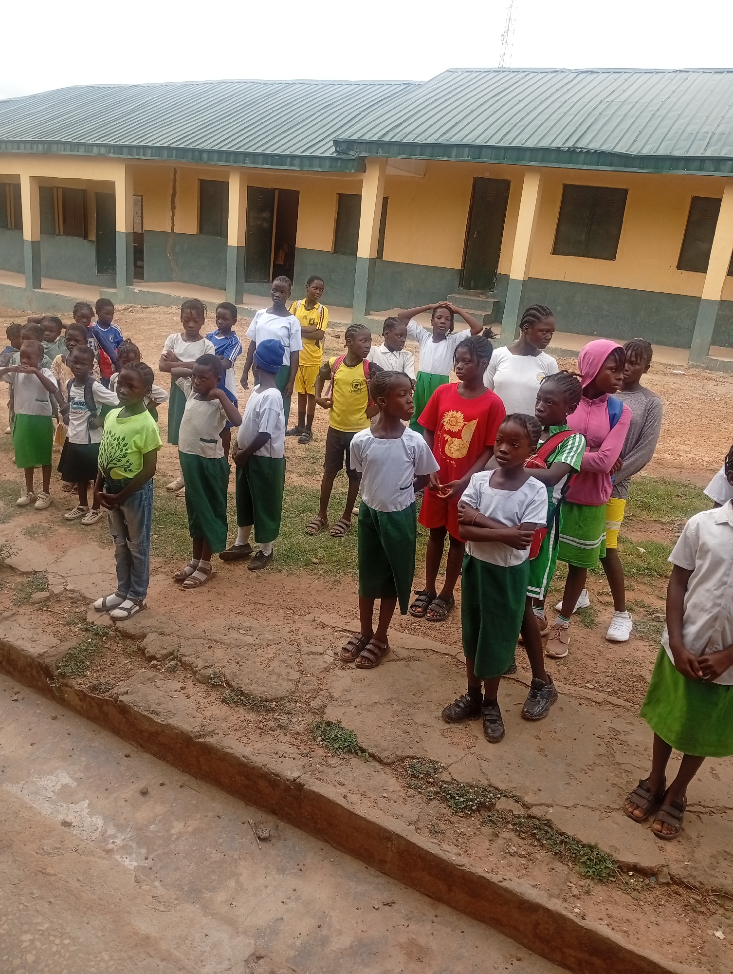 A group of approximately 20 children stand outside a single-story building with a green roof, likely a school, in an unspecified location in the city of Unknown, Unknown. The children are dressed in a variety of clothing, including school uniforms consisting of green skirts or shorts and white shirts, as well as some casual wear like t-shirts and jeans. They are arranged in a loose line, some with arms crossed, and appear to be waiting. The environment is a paved area with visible cracks and weeds growing in the crevices. The building has several windows and doors, some open.