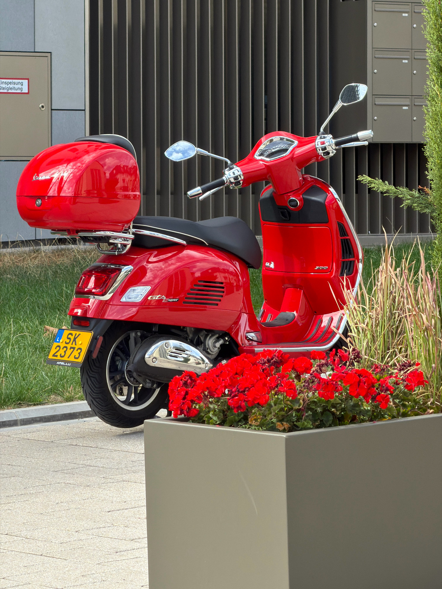 A bright red Vespa GTS Super scooter is parked on a paved area next to a planter filled with red flowers and ornamental grasses. The scooter has a black seat and a red top case. A yellow license plate reading "LSK 2373" is visible on the rear fender. In the background, a building with vertical metallic siding and a series of mailboxes can be seen. A sign with German text that reads "Einspeisung Steigleitung" is attached to the building. The scene appears to be in a contemporary urban setting, likely Esch-sur-Alzette, Luxembourg, given the license plate format and the architecture. There are no people in the image.