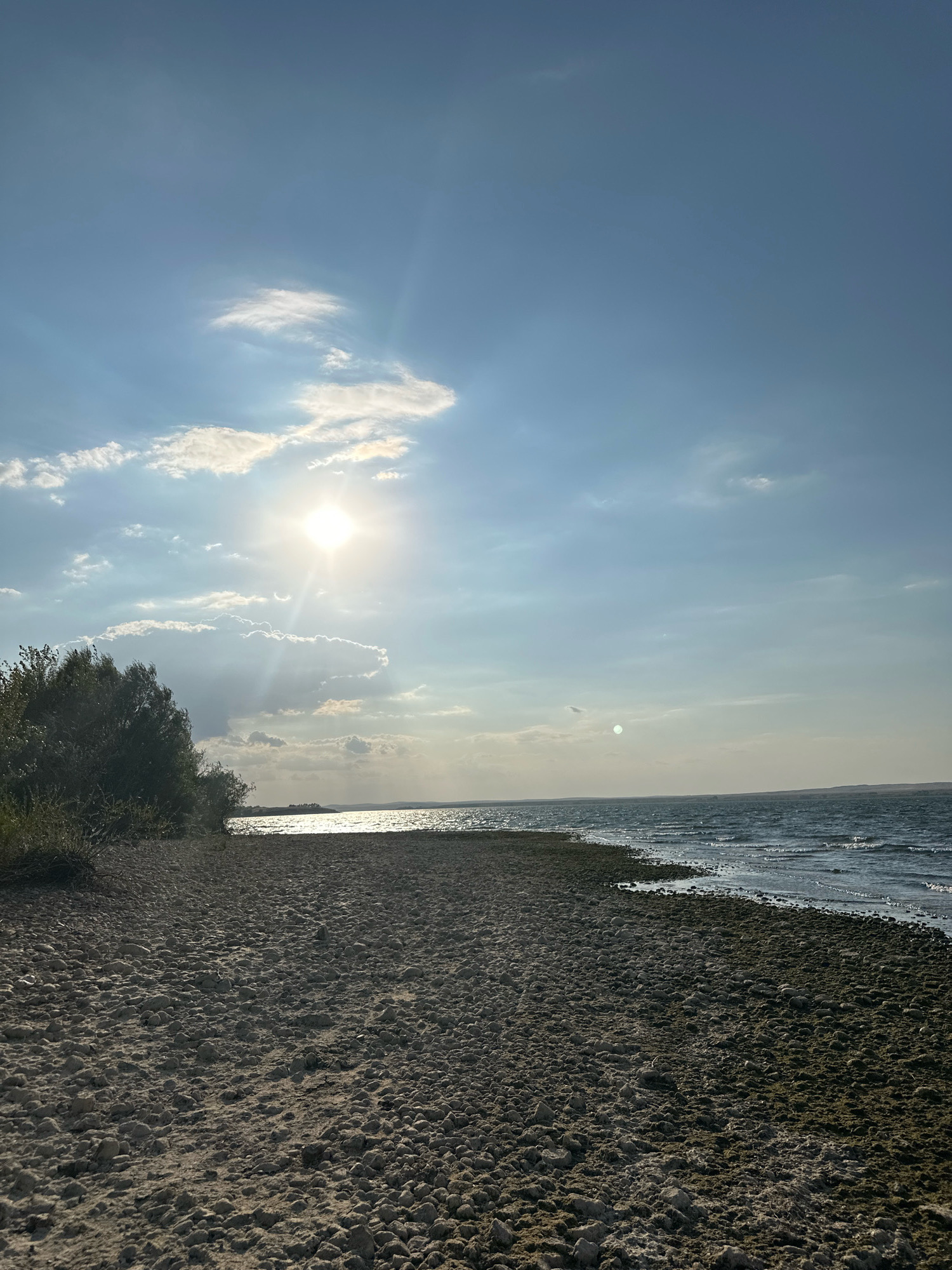 The image depicts a serene natural landscape under a partly cloudy sky. The sun, a bright orb, shines through scattered clouds, casting sunbeams across the scene. In the foreground, a coarse, pebbly beach stretches towards the water. To the left, lush green foliage forms a dense thicket. The water, a deep blue, is agitated with small waves, suggesting a gentle breeze. The horizon is marked by a distant shoreline, barely visible under the hazy sky. The overall impression is one of tranquility and natural beauty. This scene is consistent with coastal areas or large lakeshores. However, without specific landmarks, it is impossible to confirm its exact location within the city of Energetik, Russia.