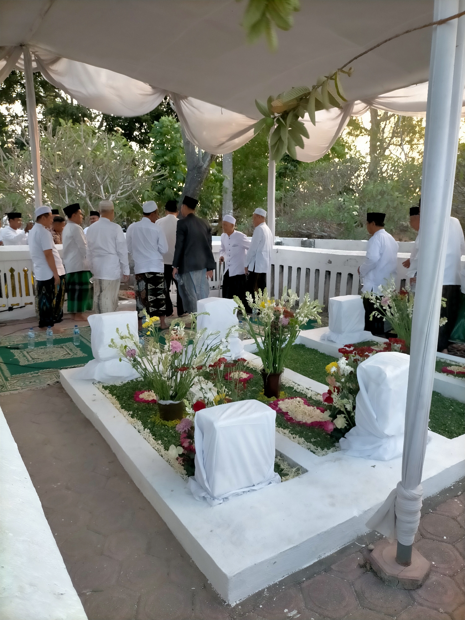 In the city of Kranji, Indonesia, a gathering of men in traditional Indonesian attire, including peci caps and sarongs, are seen around a cemetery. The environment suggests a respectful or commemorative occasion, possibly a burial or remembrance service. The men are standing and observing a row of graves, which are adorned with white cloths and flowers. The graves are arranged in a neat line, and the surrounding area is paved and covered with green carpeting. The scene is set outdoors, under a white canopy, with trees and foliage visible in the background, illuminated by what appears to be late afternoon sunlight.