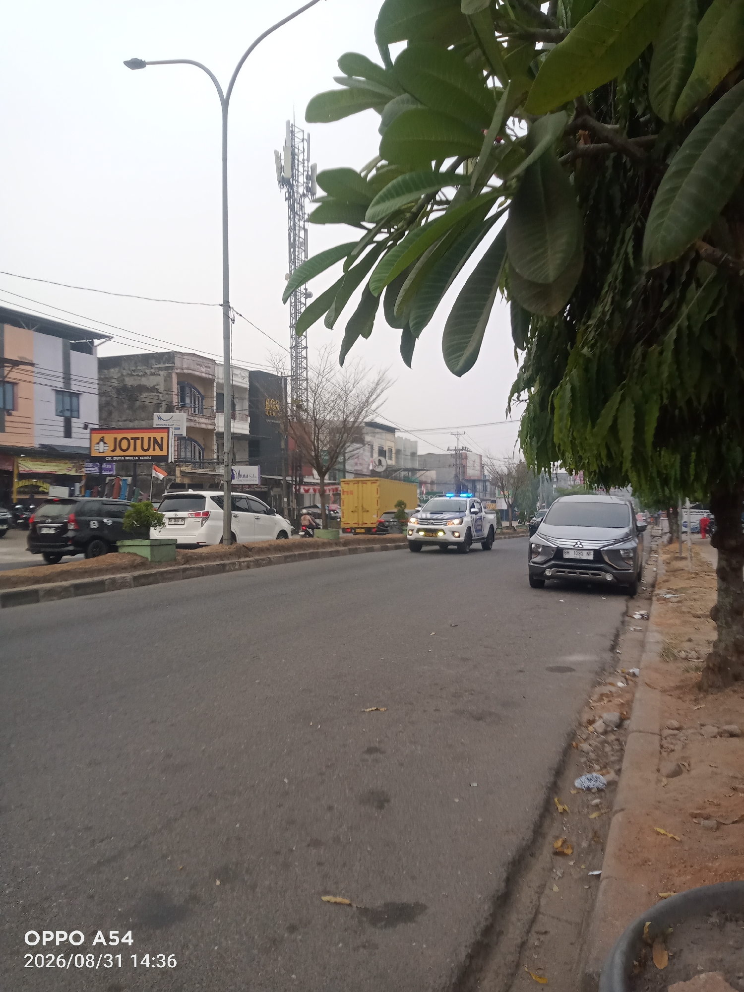 The image captures a street scene in Jambi, Indonesia, as indicated by the timestamp and the visible license plate format "BH". The foreground shows a paved road with a gray SUV parked on the right side. Further down the road, a white police car with flashing blue lights is visible, suggesting law enforcement activity. On the left side of the road, several cars are parked, including a black SUV and a white sedan. Buildings line the street, with one prominent building displaying a "JOTUN" sign. A cell tower stands in the background, and a large tree with green leaves partially obscures the view on the right. The sky is overcast, and the overall atmosphere is hazy. Debris is scattered along the curb. The timestamp on the image reads "2026/08/31 14:36".