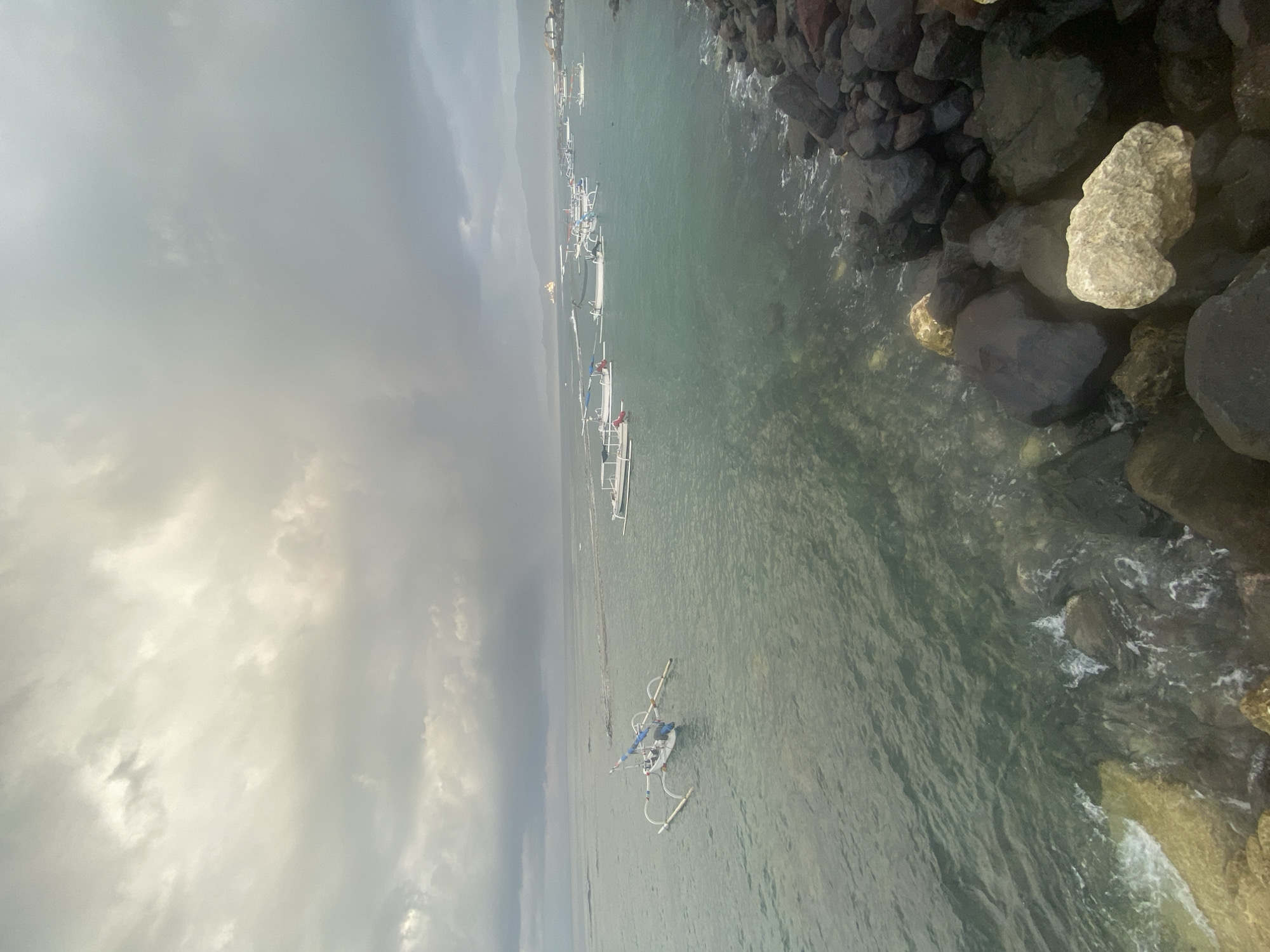 An overcast sky looms over the turquoise waters of a bay in Karangasem, Indonesia, where a fleet of traditional outrigger boats, known as jukungs, are moored near a rocky shoreline. The boats, mostly white with blue and red accents, are arranged in a line stretching into the distance, with one prominent jukung in the foreground featuring a more elaborate sail and design. The water is calm, with gentle waves lapping against the dark, rough-hewn rocks that form the immediate foreground. In the background, a hazy coastline with mountains is visible under the dramatic, cloud-laden sky. The overall scene evokes a sense of serene, yet potentially dramatic, coastal weather.