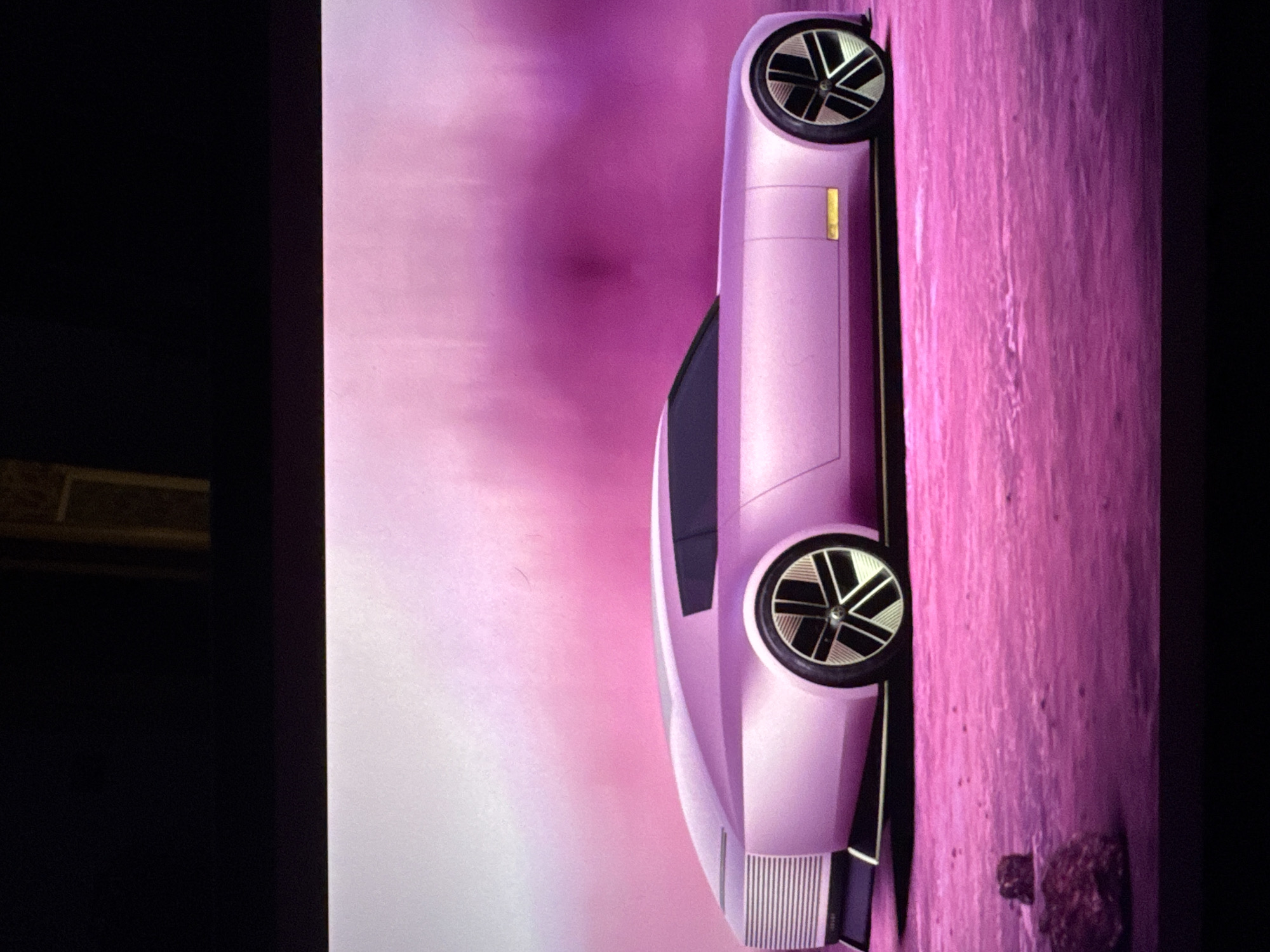 The image is a digital rendering of a futuristic concept car, presented in a profile view. The car is a light pink or lavender color, with a sleek, elongated silhouette. It features a distinctive front grille design, dark tinted windows, and large, multi-spoke wheels with a black and silver finish. The car is positioned on a pink, textured surface that resembles a desert or barren alien landscape, with a few dark-colored rocks scattered in the foreground. The background is a gradient of soft pink and white, suggesting a sky or atmospheric effect. There is no indication of the city of Sant'Angelo di Piove di Sacco, Italy, in this image.