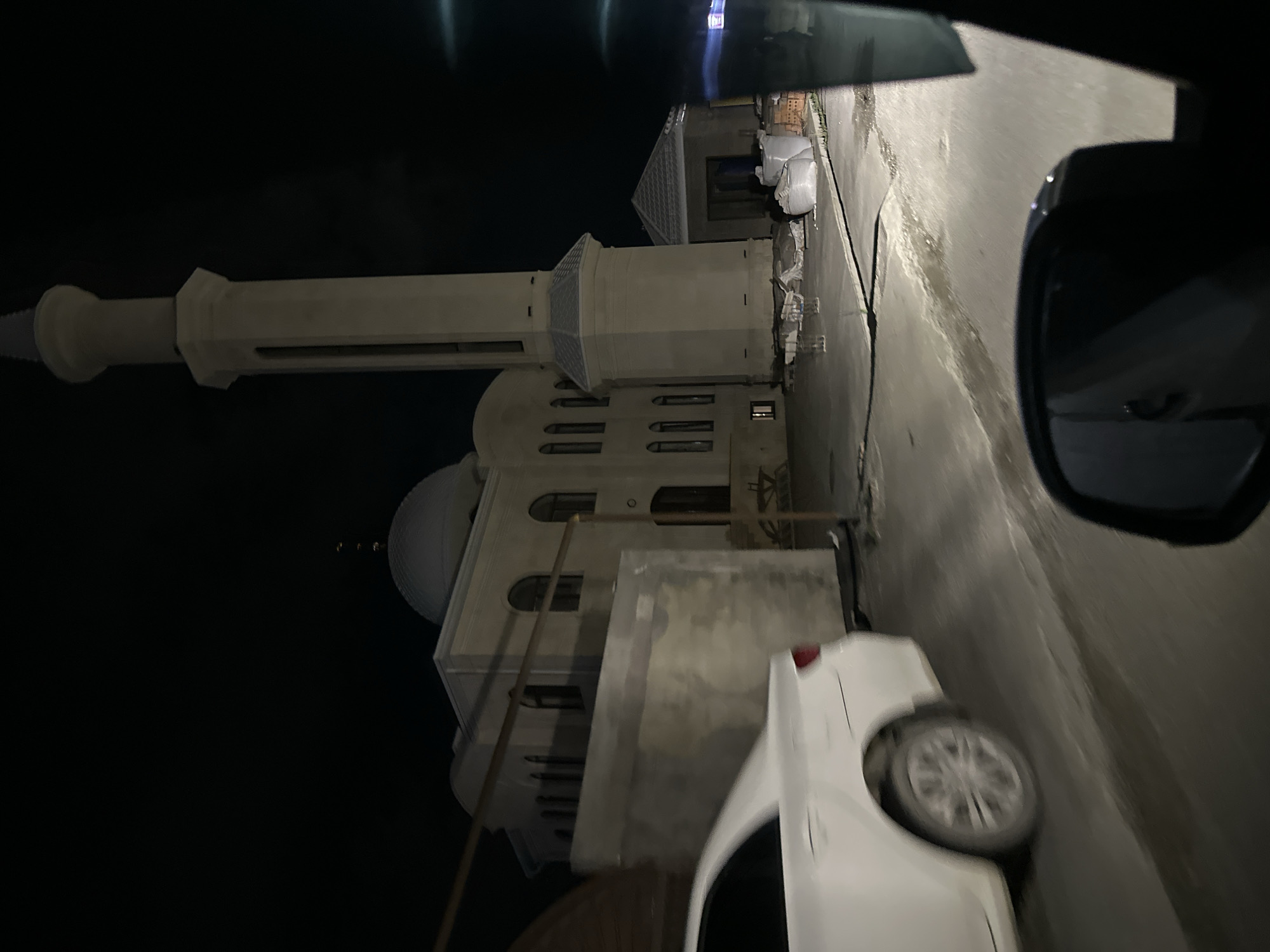 In Miatli, Russia, a white sedan is captured in motion at night, its rear section visible in the foreground, suggesting a forward-moving perspective from within another vehicle. The road appears wet or reflective, illuminated by artificial light sources. Adjacent to the road, a light-colored mosque stands prominently, featuring a large dome and a tall, slender minaret. The mosque is illuminated, highlighting its architectural details and the surrounding construction materials, including sacks of what appear to be cement or sand and stacks of bricks. The environment is dark, indicating nighttime, with the sky a deep black.