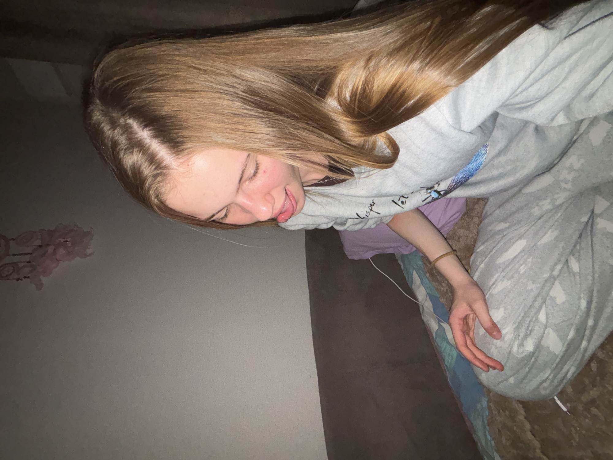 A young woman with long, light brown hair is sitting on a couch, with her head tilted down and her tongue sticking out. She is wearing a grey sweatshirt with a design on the front and grey patterned sweatpants. Her right hand is resting on her lap. The couch is brown, and there is a purple pillow behind her. The background is a plain wall with a dreamcatcher hanging on it. The scene appears to be indoors, in a residential setting. The location is not specified in the image.