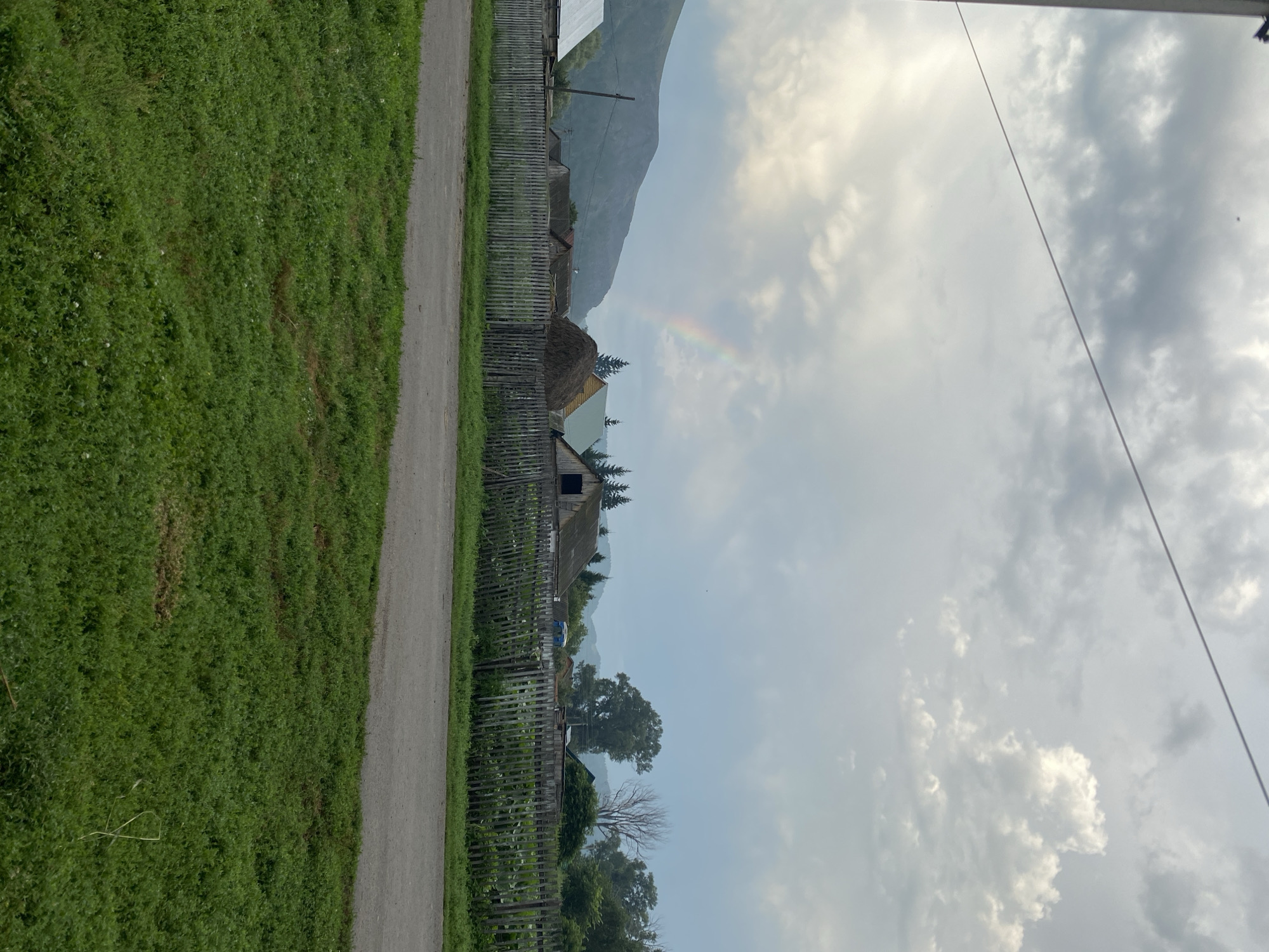 In Maloarslangulovo, Russia, a rural landscape unfolds under a cloudy sky with a faint rainbow arcing over a village. A wooden fence borders a paved road, with a strip of green grass in the foreground. Behind the fence, several wooden houses with weathered roofs are visible, one topped with a large haystack. In the distance, a mountain range is shrouded in haze, and evergreen trees dot the landscape. The scene is peaceful and serene, capturing the quiet beauty of the Russian countryside.