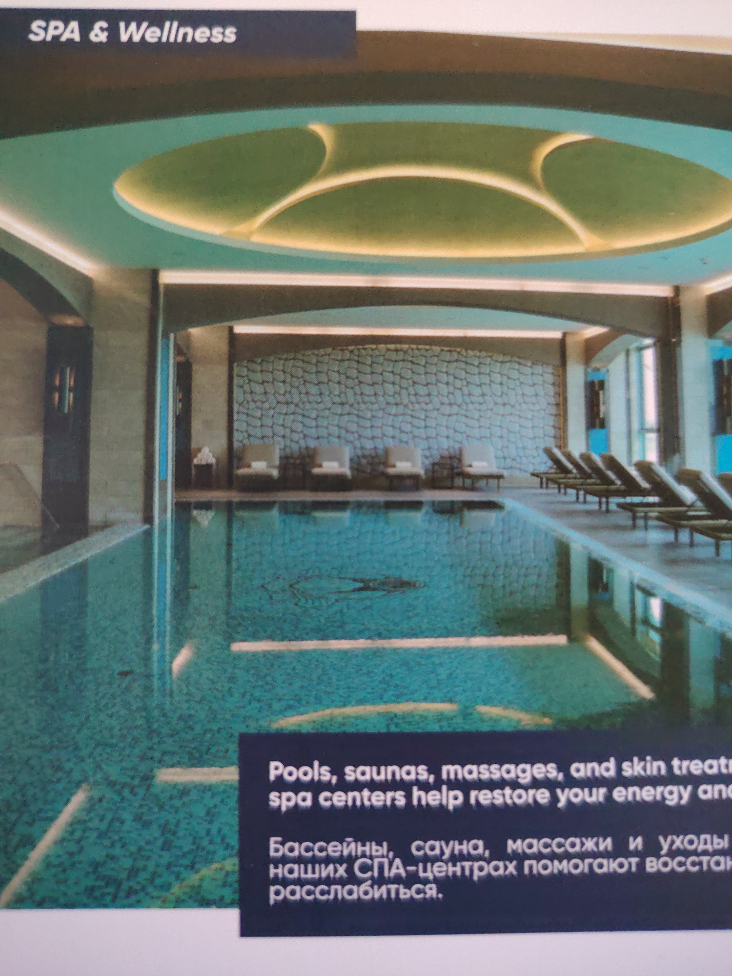 The image depicts a luxurious indoor swimming pool area within a spa and wellness center. The pool is illuminated by overhead lighting and features a mosaic tile bottom. Around the pool, there are several lounge chairs and some smaller chairs with towels. The walls are decorated with a textured pattern, and there are modern light fixtures. The ceiling has an interesting design with curved elements and integrated lighting. A dark blue sign in the foreground displays text in both English and Russian, describing the spa services offered, including "Pools, saunas, massages, and skin treatments help restore your energy and relax." The image does not contain any information that indicates the location is in Bahshitpa, Uzbekistan.