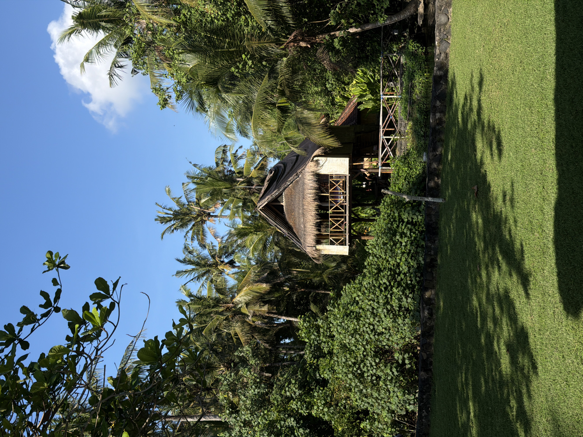 The image shows a thatched-roof structure situated among palm trees and lush vegetation in Karangasem, Indonesia. The sky is blue with a few scattered clouds. The ground to the side is covered in green grass. The structure is elevated with an open design featuring a bamboo railing. The photograph has been rotated to a vertical position.