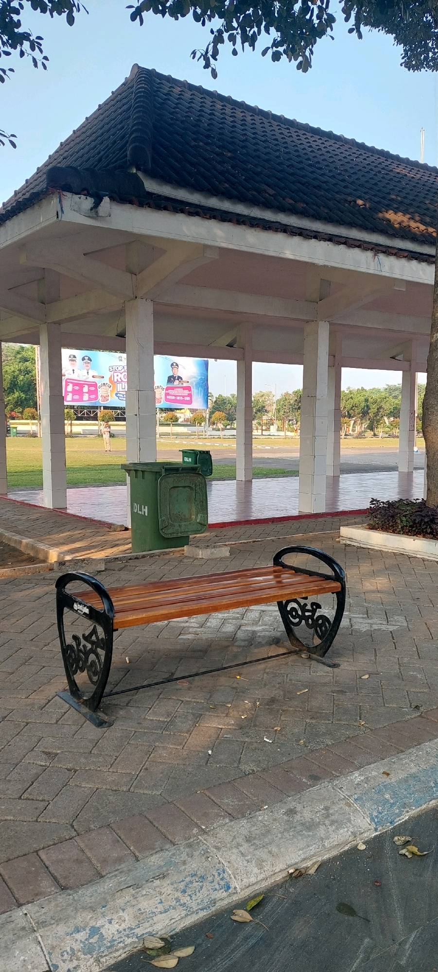 The image depicts an outdoor scene at the Lumajang City Square in Lumajang, Indonesia. In the foreground, a wooden bench with decorative black metal armrests, featuring bicycle motifs, is placed on a paved walkway made of interlocking bricks. Beyond the bench, two green trash bins with "DLH" (Dinas Lingkungan Hidup, or Environmental Agency) labels are visible. In the background, a covered pavilion with a tiled roof and white pillars stands open, revealing billboards behind it. The pavilion is surrounded by a manicured lawn and trees, and a paved area with red borders. The overall environment suggests a public park or recreational area. The sky is clear and blue, indicating daytime.