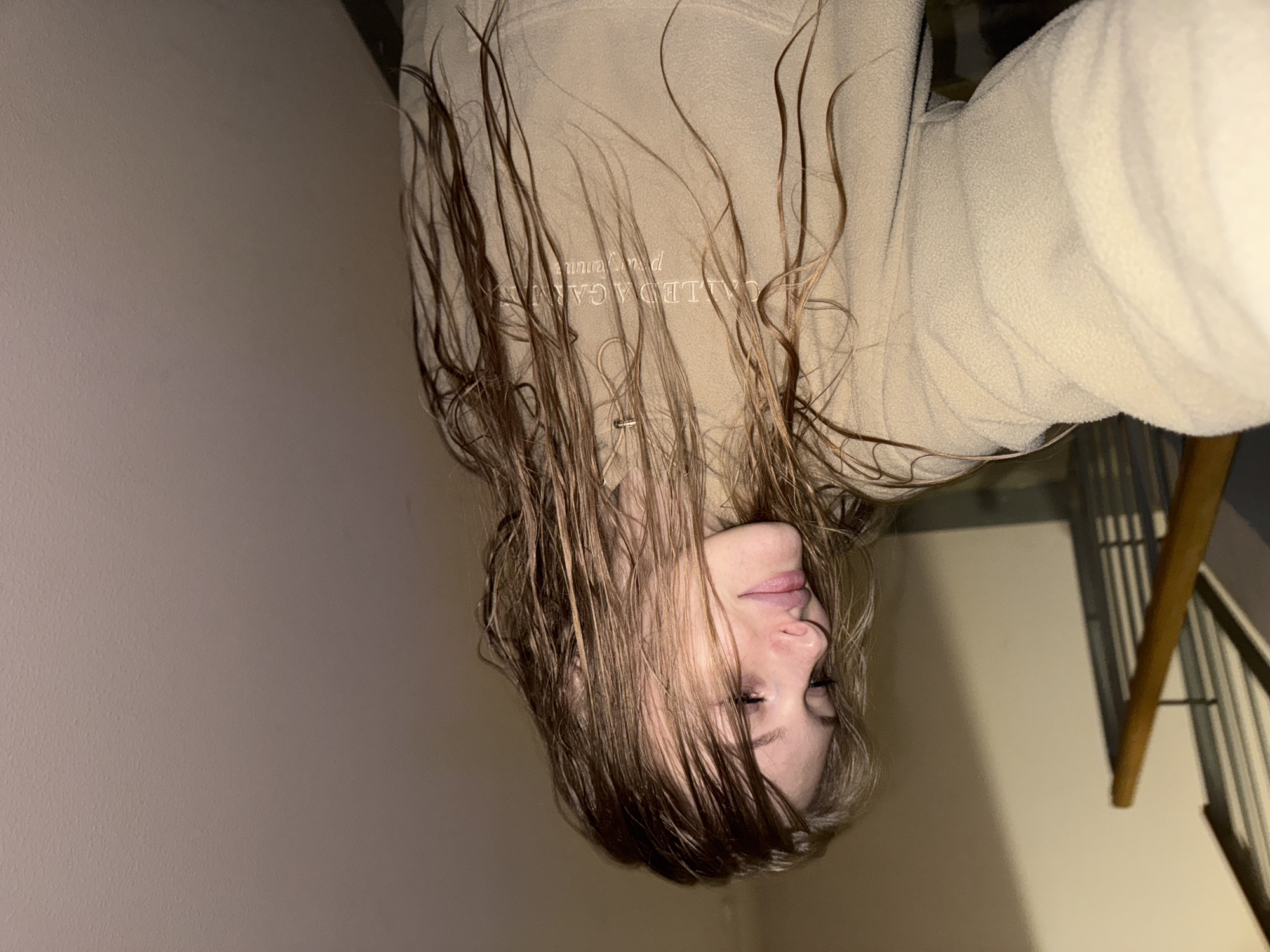 A young female with light brown, wavy hair is shown from the chest up, angled downwards. She is wearing a light-colored fleece jacket. The background is a textured, light-colored wall and a railing of a staircase. There is text embroidered on her jacket. The overall lighting suggests an indoor environment, possibly a building or stairwell. The specific location in Kondratovo, Russia, is not visually evident in the image.