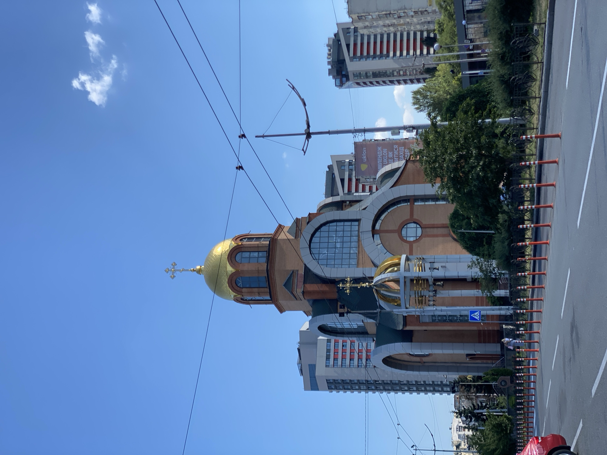 The image depicts a bright, clear day in a city, likely in Eastern Europe due to the architectural style and script on the billboard. In the foreground, a multi-lane road with white lane markings is visible, with a red car on the left edge and an array of orange and white traffic cones lining the roadside. A black metal fence runs along the sidewalk, behind which is a row of trees and bushes.
The focal point of the image is a large, ornate church with a prominent golden dome topped with a cross. The church is constructed of brick and features arched windows and decorative architectural elements. To the left and right of the church, modern high-rise buildings rise into the blue sky. A billboard with Cyrillic script and the logo "Uklon" is visible on a pole between the church and one of the modern buildings.
Several people are visible walking on the sidewalk near the church, and a few birds are perched on overhead power lines. The sky is a vibrant blue with a few wispy white clouds. The overall scene suggests a busy urban environment where religious and modern architecture coexist.
