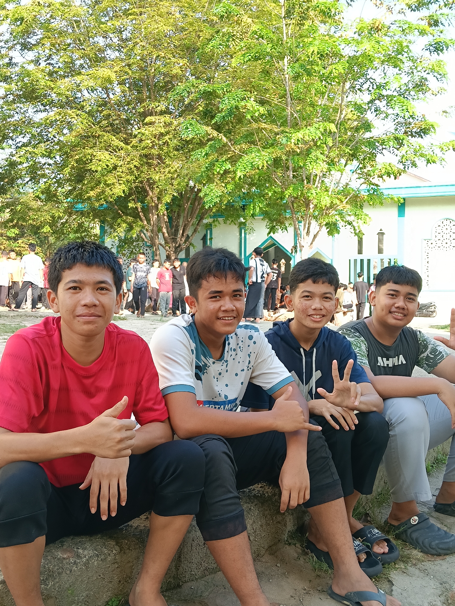 Four young males are seated in the foreground, posing cheerfully for a photo. The individual on the far left, in a red t-shirt and black cropped pants, gives a thumbs-up. Next to him, a male in a white and blue patterned t-shirt and black cropped pants smiles. The third male, wearing a dark blue hooded top and black cropped pants, holds up a peace sign. The fourth, on the far right, in a dark green patterned t-shirt and light grey pants, also smiles broadly. All four appear happy and relaxed.
In the background, a crowd of other people, predominantly young males, are visible standing and moving around, suggesting a gathering or event. Behind them stands a building with light-colored walls and distinctive green or teal trim around its windows and roofline, which appears to be a mosque or an Islamic educational institution. A large tree with dense green leaves dominates the upper portion of the scene, providing shade.
The photo was taken outdoors in bright daylight, indicated by the clear sky and the shadows cast by the subjects and tree, suggesting a morning or late afternoon setting. Visible text on clothing includes a partial brand name "PERTAMIN" (likely "PERTAMINA") on the white and blue t-shirt and "AHLIA" on the dark green t-shirt. One pair of sandals has a design resembling Crocs. The overall mood is casual and friendly.