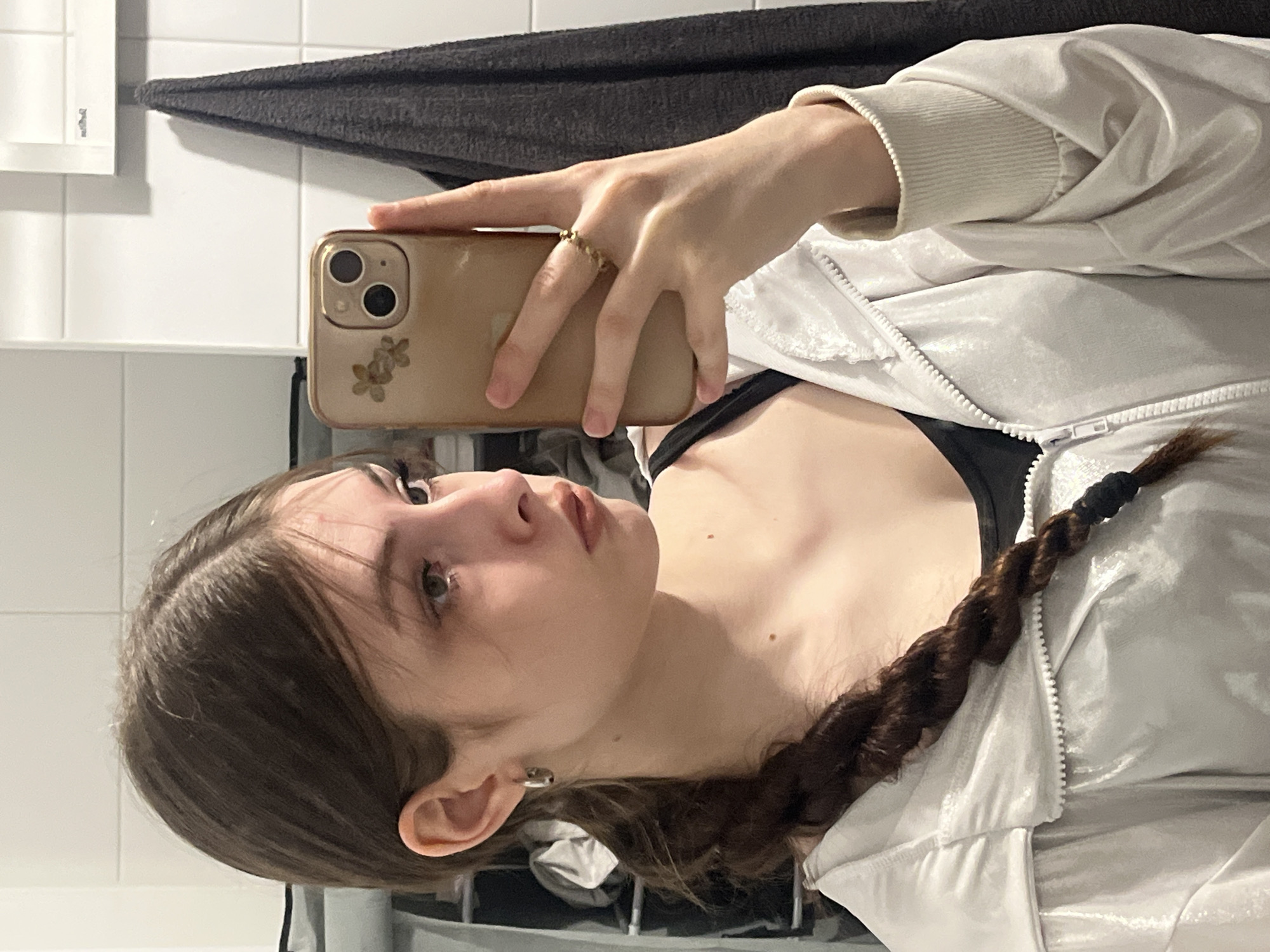 The image, oriented vertically with the person's head to the right, captures a young person taking a mirror selfie. The subject is a young individual with fair skin and dark brown hair, styled in a long braid that extends over their chest and shoulder. Several small moles are visible on their neck and upper chest.
They are wearing a light-colored, possibly silver or white, shiny puffer jacket with a visible zipper, layered over a black top. On their right ring finger, they wear a gold-colored ring with small beaded details. Their right hand holds a light gold-colored smartphone, possibly an iPhone, with a visible dual-camera system and a floral sticker on the back case. A silver-toned hoop earring is worn in their left ear.
The background consists of white square tiles, strongly suggesting a bathroom setting. A dark grey or black towel is draped in the upper left portion of the background, and a white rectangular fixture is partially visible on the far left. The person's gaze is directed upwards and slightly to the right, appearing to look at their reflection or the phone screen while taking the picture. The lighting is artificial, consistent with an indoor environment. No legible text is visible in the image.