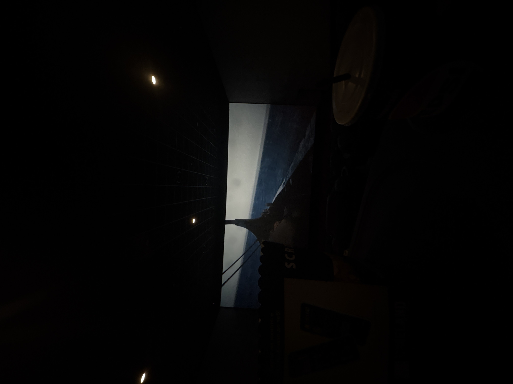 The image displays the interior of a darkened cinema hall in Plovdiv, Bulgaria, viewed from a spectator's perspective. A large screen at the center projects a film scene depicting a tall, dark structure on the left against a muted blue-gray background, suggestive of a maritime or stormy environment. In the immediate foreground on the left, a dark popcorn bucket, partially obscuring the screen, shows the fragmented text "SCRA". Below it, a portion of a light-colored rectangular object is visible. On the immediate right, a beverage cup with a light beige lid and a dark straw is positioned. The cinema ceiling, dark and featuring a grid pattern, has several small, illuminated circular lights; two are distinctly visible in the upper left and upper right areas of the frame. No individuals are discernible within the visible scene.
