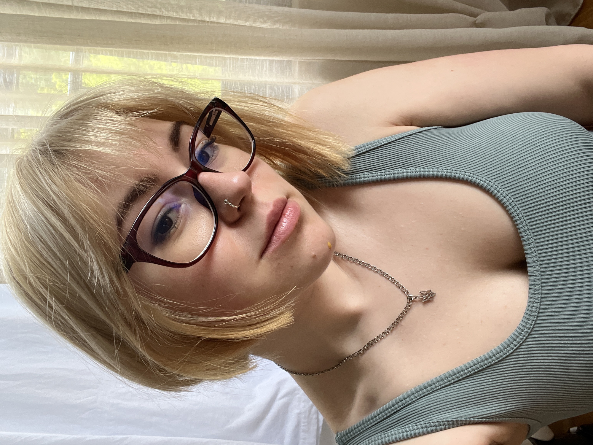 A woman with blonde hair and glasses is lying on a white surface. She is wearing a ribbed tank top and a silver necklace with a star pendant. She has a nose piercing. The background is a window with light filtering through curtains. The location in the city of Boreti, Montenegro is not discernible in the image.