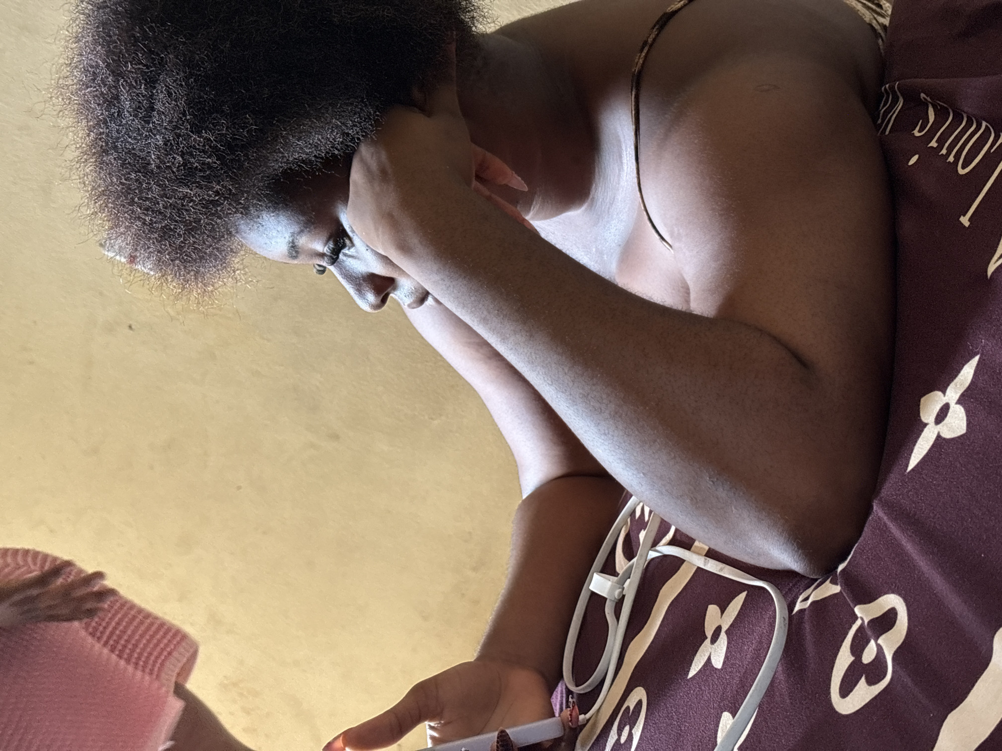 The image, rotated on its side, captures a close-up, low-angle view of an individual, likely female, with dark skin and voluminous natural, curly dark hair. The person is positioned indoors, leaning forward with a downward gaze, suggesting focus or introspection. Their face, seen in profile, shows a concentrated expression.
The individual appears to be wearing a sleeveless garment or is bare-chested, with a thin strap visible over their shoulder. One arm is bent, with the hand displaying long, light-colored manicured nails, resting gently near their jaw or neck. The other hand holds a slender, clear object, possibly an e-cigarette or vape pen, with red nail polish visible on the fingertips. A white cord, likely from headphones or a charging cable, is draped over their lower arm and the adjacent patterned fabric.
To the right, a dark brown textile, possibly a blanket or bed sheet, is visible. It features a repeating pattern of light brown or cream motifs resembling clover leaves or flowers, along with partial text that clearly spells "Louis V," indicating the Louis Vuitton brand. In the bottom left, a small section of a pink, ribbed fabric and a hand are visible, hinting at the presence of another person or item.
The background consists of a plain, light yellow or beige wall or surface. The lighting appears to be natural, suggesting the photo was taken during the daytime. The scene conveys a personal and quiet moment in an indoor setting, located in Ikot Ekpene, Nigeria.