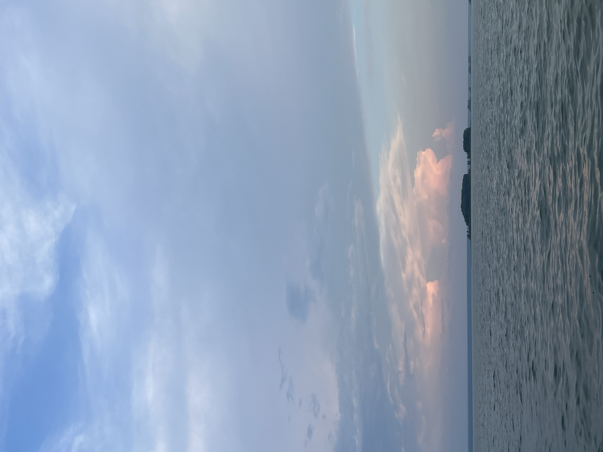 The image shows a wide expanse of water under a cloudy sky. In the distance, there are several small islands covered in trees. The sky is filled with various shades of blue and gray clouds, with some areas tinged with pink, suggesting either sunrise or sunset. The water is choppy, with small waves creating ripples on the surface. The horizon is clear, with a faint line of land visible in the far distance. The scene is calm and serene, evoking a sense of vastness and natural beauty. The location is stated to be in Novopichugovo, Russia. There are no people or specific objects of interest visible in the image.
