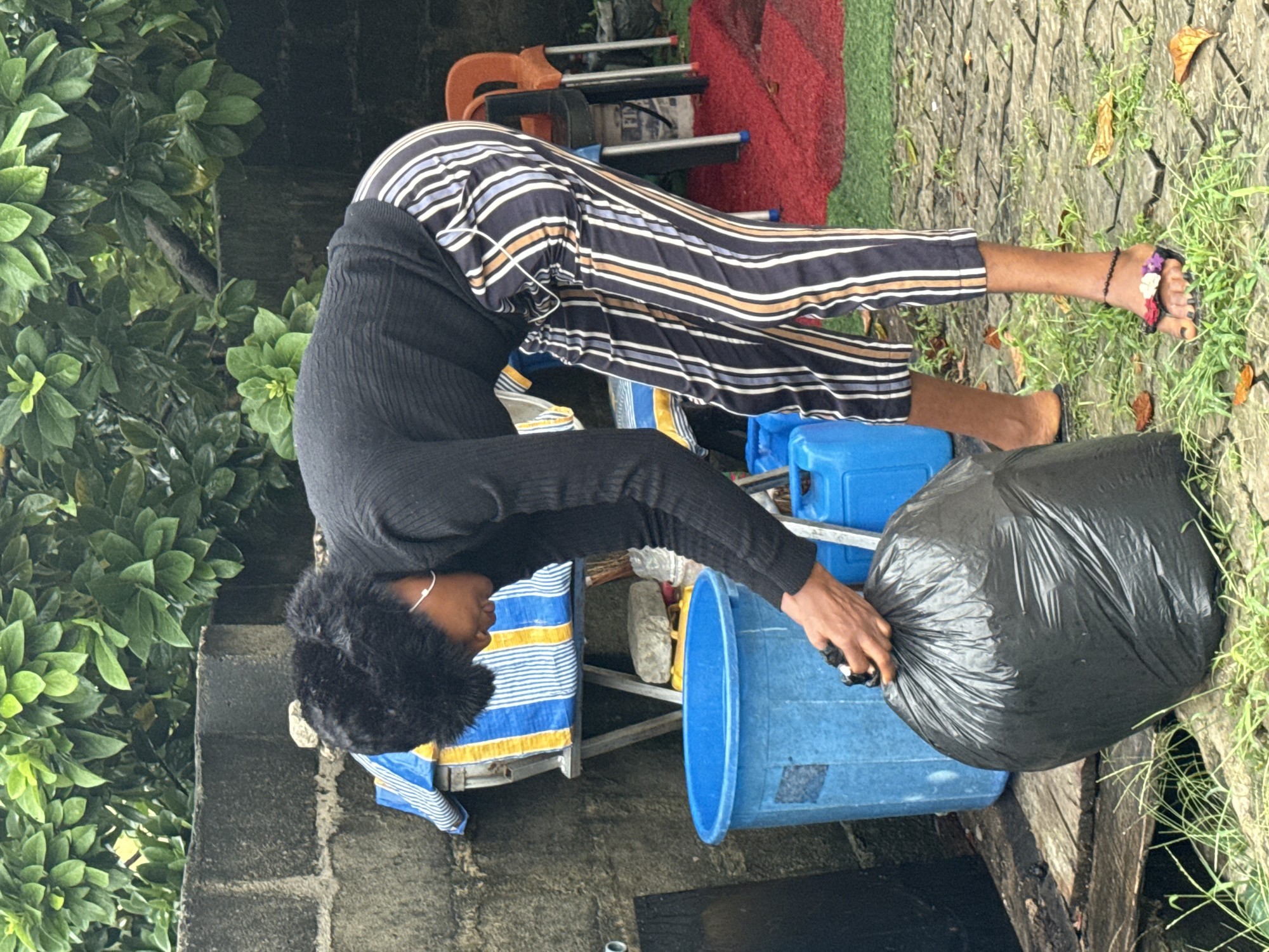 A person is bending over to place a black plastic trash bag in a blue plastic bin outdoors. The person is wearing a black long-sleeved shirt, striped pants, and sandals with floral embellishments on one foot. The bin is positioned near a concrete wall with green foliage and a paved area with scattered leaves. Other items, including blue plastic containers and a folded metal frame with striped fabric, are nearby. The location is suggested to be in the city of Ado, Nigeria.