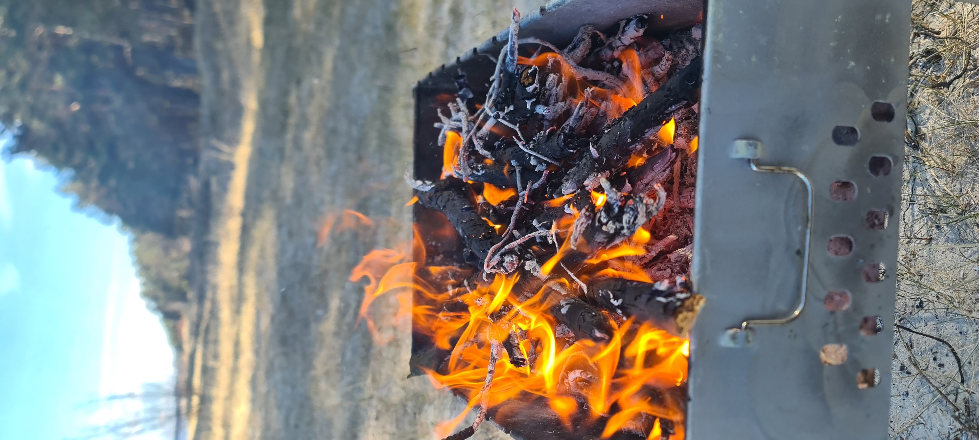 The image shows a metal grill filled with burning wood and branches. The flames are orange and yellow, and the wood is black and charred. The grill is sitting on the ground, with grass and trees visible in the background. The location appears to be outdoors, possibly in Zīvartiņš, Latvia. The image primarily captures a fire burning in a portable grill, in an outdoor setting.