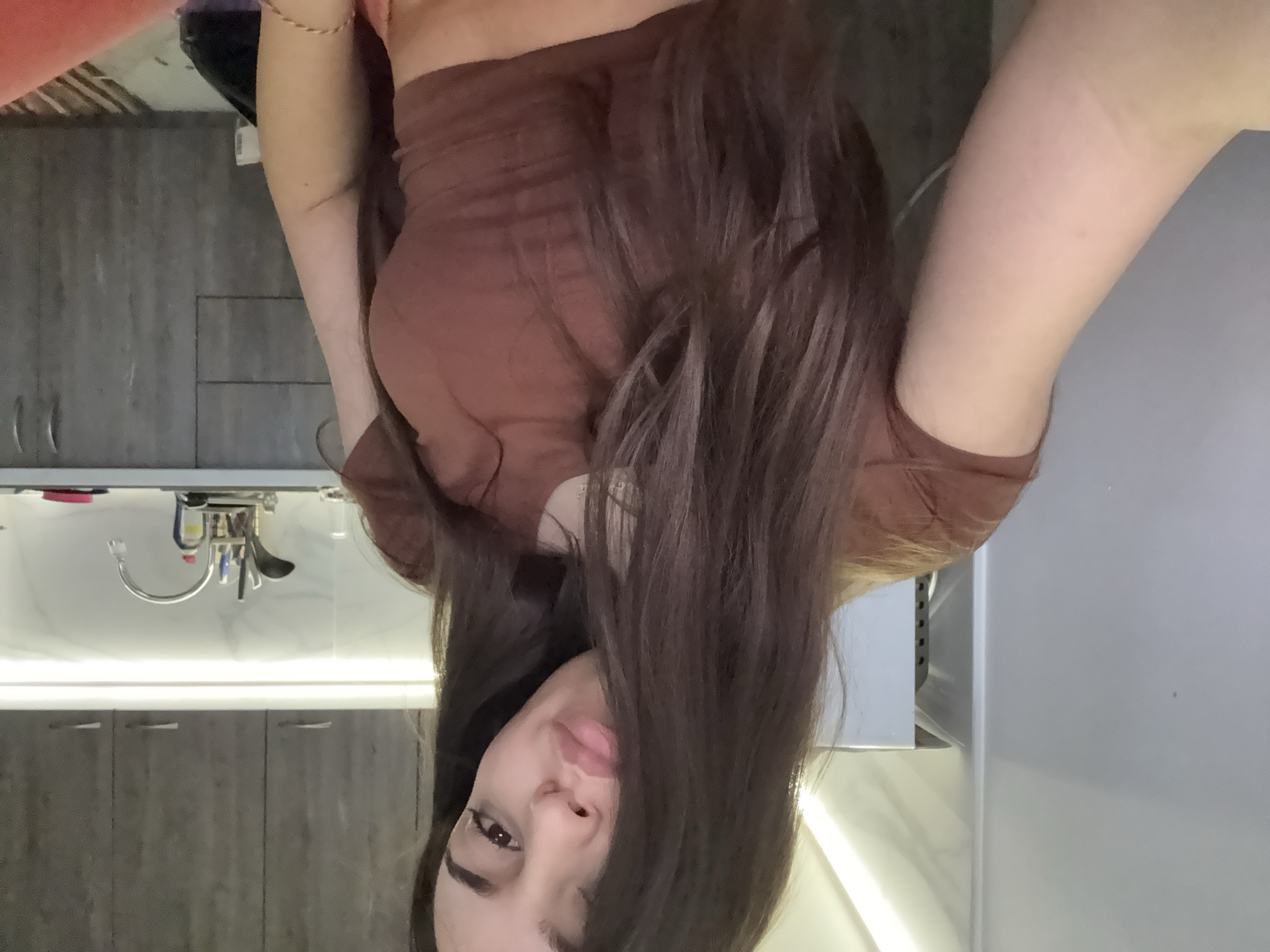 A woman with long, dark hair is positioned in the center of the frame, wearing a brown top. The background suggests a kitchen or bathroom setting. The sink with a faucet and cabinets are visible. The wall and additional surfaces are white. There is no information provided to indicate this is in Plovdiv, Bulgaria.