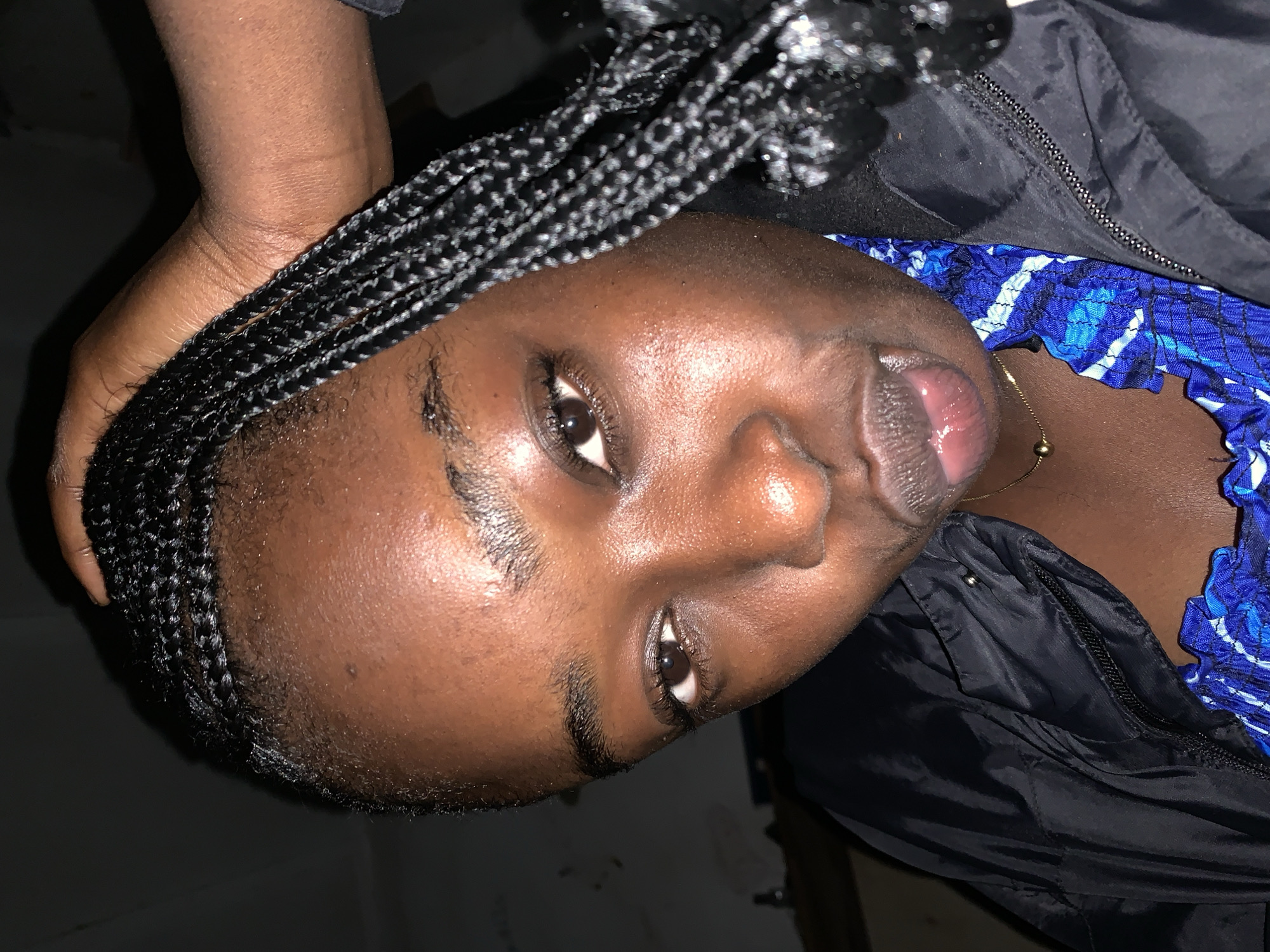 The image is a close-up, low-angle selfie of a young Black woman. She has dark skin, braided hair, and is wearing a black jacket over a blue and white patterned top. A delicate gold necklace with a single bead is visible. Her left hand is raised, touching the top of her head and pulling some of her braids forward. The background is dark and indistinct, suggesting an indoor setting, possibly at night. The scene is located in Rafin Par, Nigeria.