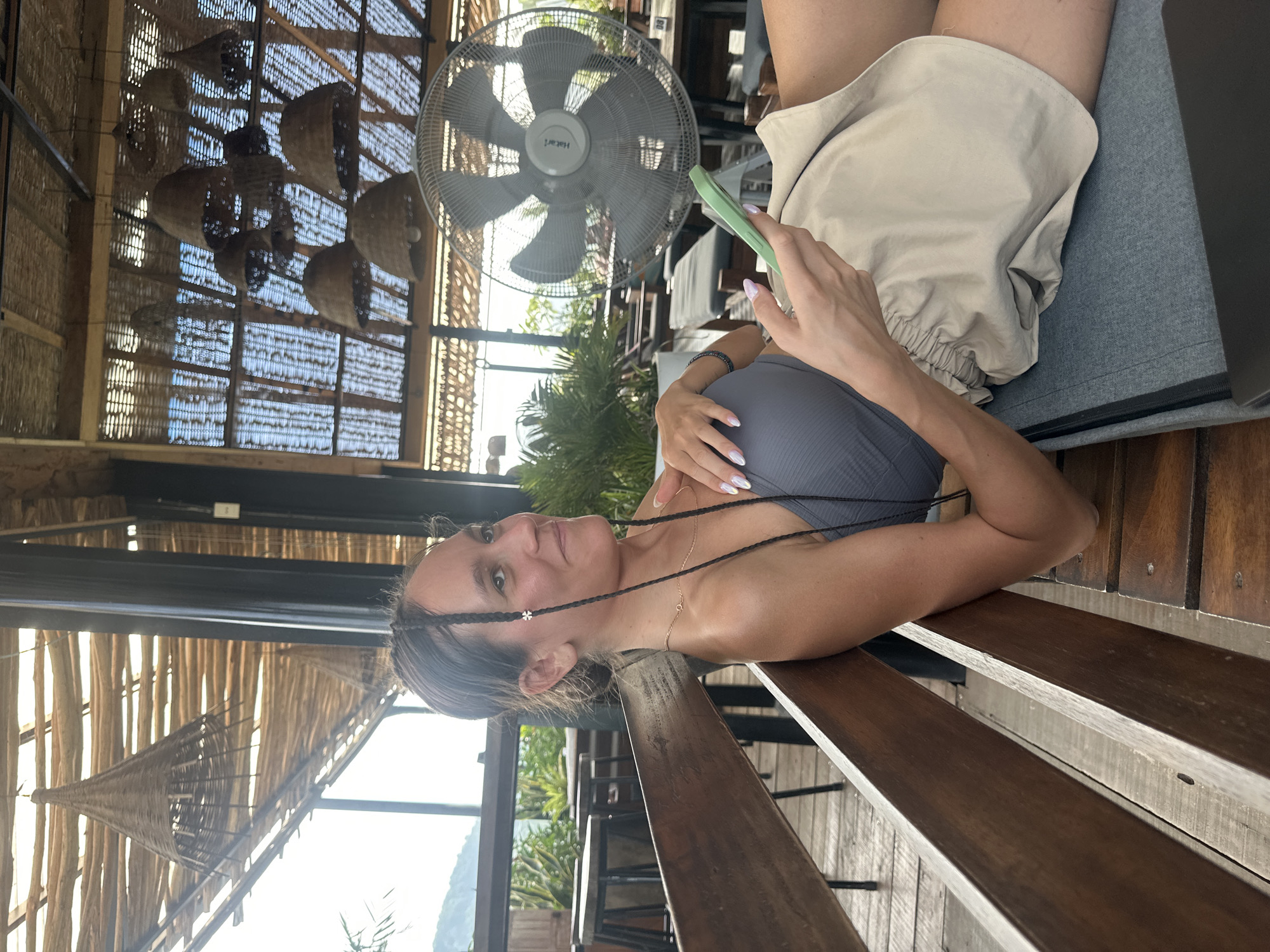 An eye-level shot captures a woman relaxing in what appears to be a semi-open, rustic cafe or lounge area in Hà Giang 1 Ward, Vietnam. She is positioned reclining or sitting comfortably on a bench, looking towards the left of the frame with a gentle smile, suggesting contentment.
The woman has dark hair styled with thin braids, and she wears a grey ribbed top and light-colored, possibly beige or khaki, shorts. A delicate gold chain necklace is visible around her neck, and she has a dark bracelet on her left wrist. Her fingernails are long and manicured with a light, iridescent polish. In her right hand, she holds a light green mobile phone. Next to her leg, a dark object, possibly a laptop, is partially visible.
The setting features natural materials, including a ceiling constructed from woven straw or bamboo panels with exposed wooden beams. Several decorative woven basket-like or conical lamps hang from the ceiling. A large grey standing fan, with the brand name "Hatari" visible on its center, is positioned in the upper center of the frame, suggesting a warm climate.
Through the open structure of the venue, bright daylight indicates it is daytime, with clear skies. Lush green foliage is visible in the mid-ground, and distant mountains or hills can be seen beyond, reinforcing the scenic, natural environment of the location. The furniture consists of dark wooden benches and tables.