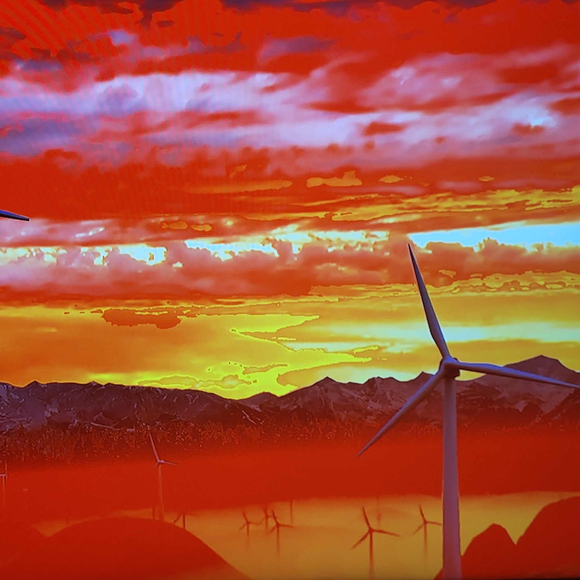 The image depicts a sunset landscape with wind turbines in the foreground. The sky is filled with red and orange hues, transitioning to yellow closer to the horizon. Mountains are visible in the midground. Several wind turbines are scattered across the scene, with one prominently positioned in the right foreground. Reflections of the turbines are visible on what appears to be a body of water. There is no evidence to place this image in Yerevan, Armenia. The location is unknown.