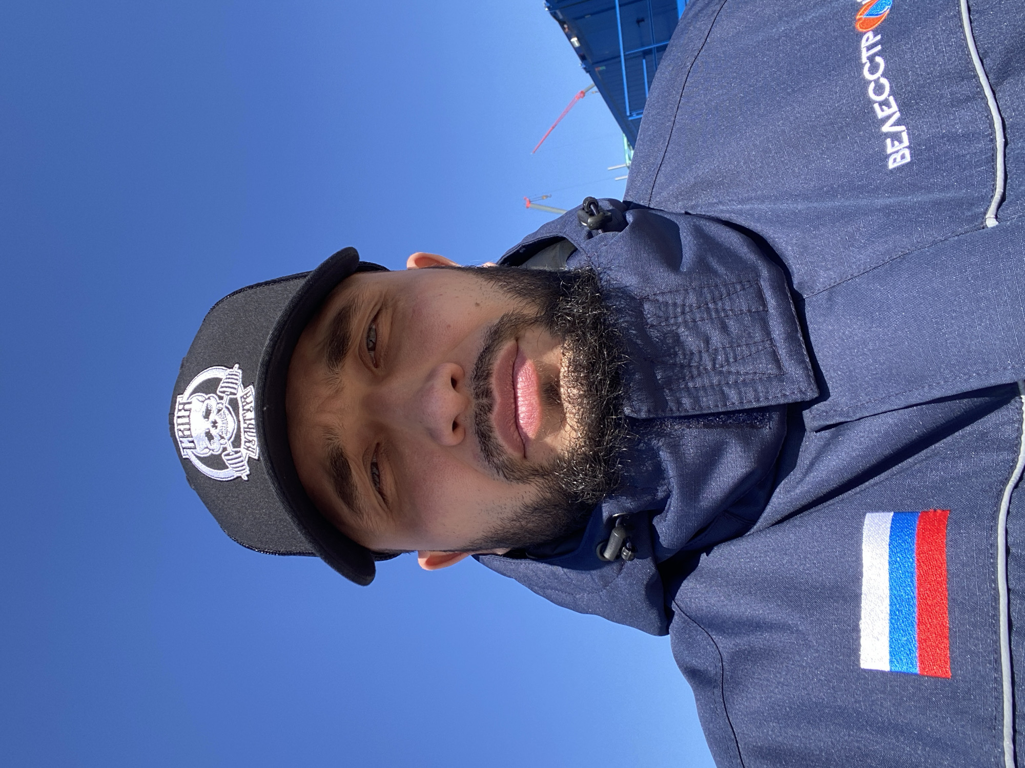 A man is depicted in a rotated portrait, likely a selfie, under a clear, bright blue sky in what is identified as Luzhitsy, Russia. He looks directly at the camera with a neutral expression. He has a dark beard and mustache.
He is wearing a dark blue jacket, which appears to be made of a textured, possibly work-grade, fabric. On the jacket's sleeve, there is an embroidered patch of the Russian flag, featuring vertical stripes of white, blue, and red. On the upper chest or shoulder area of the jacket, white Cyrillic text is embroidered, spelling out "ВЕЛЕССТР" (VELESSTR).
He also wears a black baseball cap with a white embroidered logo on the front. The logo depicts a skull design incorporating barbells, and the word "BARBELL" is visible.
The background consists primarily of an expansive, cloudless blue sky, indicating sunny weather during daytime. In the upper right quadrant of the image, a large blue structure, possibly scaffolding or part of an industrial building or ship, is partially visible against the sky, suggesting an outdoor work environment. The strong, direct lighting suggests it is mid-day.