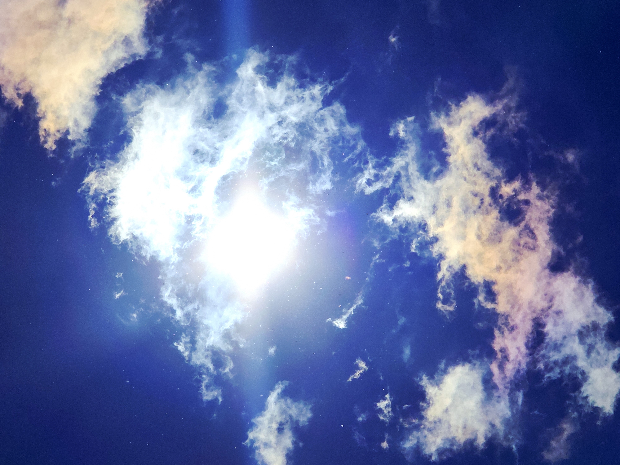 The image displays a vibrant and dynamic sky scene, dominated by a deep blue background punctuated by bright clouds and an intense light source. A powerful, brilliant light, presumably the sun, is positioned centrally in the frame, causing significant lens flare that radiates across the image.
Clouds are scattered throughout, presenting a varied appearance. Many are brilliant white, intensely illuminated by the light source, appearing bright and ethereal. Other cloud formations, particularly in the upper left and lower right sections, display warm tones, ranging from pale yellows and oranges to soft pinks and purples. These colorful clouds contrast sharply with the deep blue sky, suggesting the quality of light often seen during sunrise, sunset, or specific atmospheric conditions during the day. The clouds vary in density, from thin, wispy streaks to more substantial, fluffy clusters.
The sky itself is a rich, dark blue, providing a striking backdrop to the illuminated clouds and the bright sun. Numerous tiny white specks, possibly dust particles or atmospheric reflections, are visible scattered across the darker areas of the sky, adding a subtle texture.
No people, man-made objects, or specific geographical location cues are visible. The image focuses entirely on the natural phenomenon of clouds and sunlight. There is no visible text. The scene evokes a sense of natural beauty, vastness, and the dramatic interplay of light and atmosphere, likely captured during daytime.