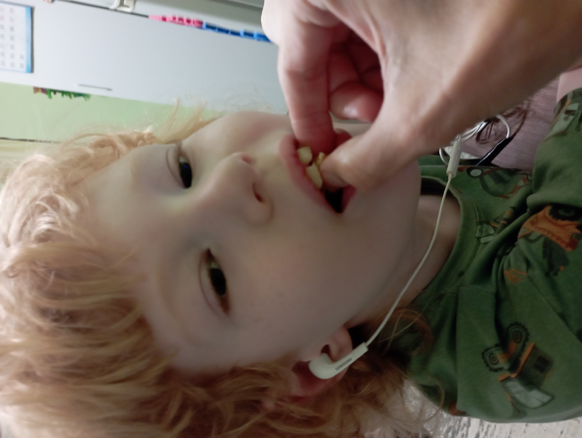 A close-up, eye-level shot shows a young child with blonde, curly hair wearing a green shirt with construction vehicle motifs. The child has white earbuds in their left ear and is being fed a small, light-colored food item by an adult's finger into their open mouth. The child's mouth is slightly agape, revealing their teeth, and they appear to be chewing. The background is blurred, but a white cabinet and a calendar are visible on the right. The scene is indoors, in what appears to be a residential setting. No specific location within the city of Blagoveshchensk, Russia can be identified from the provided image.